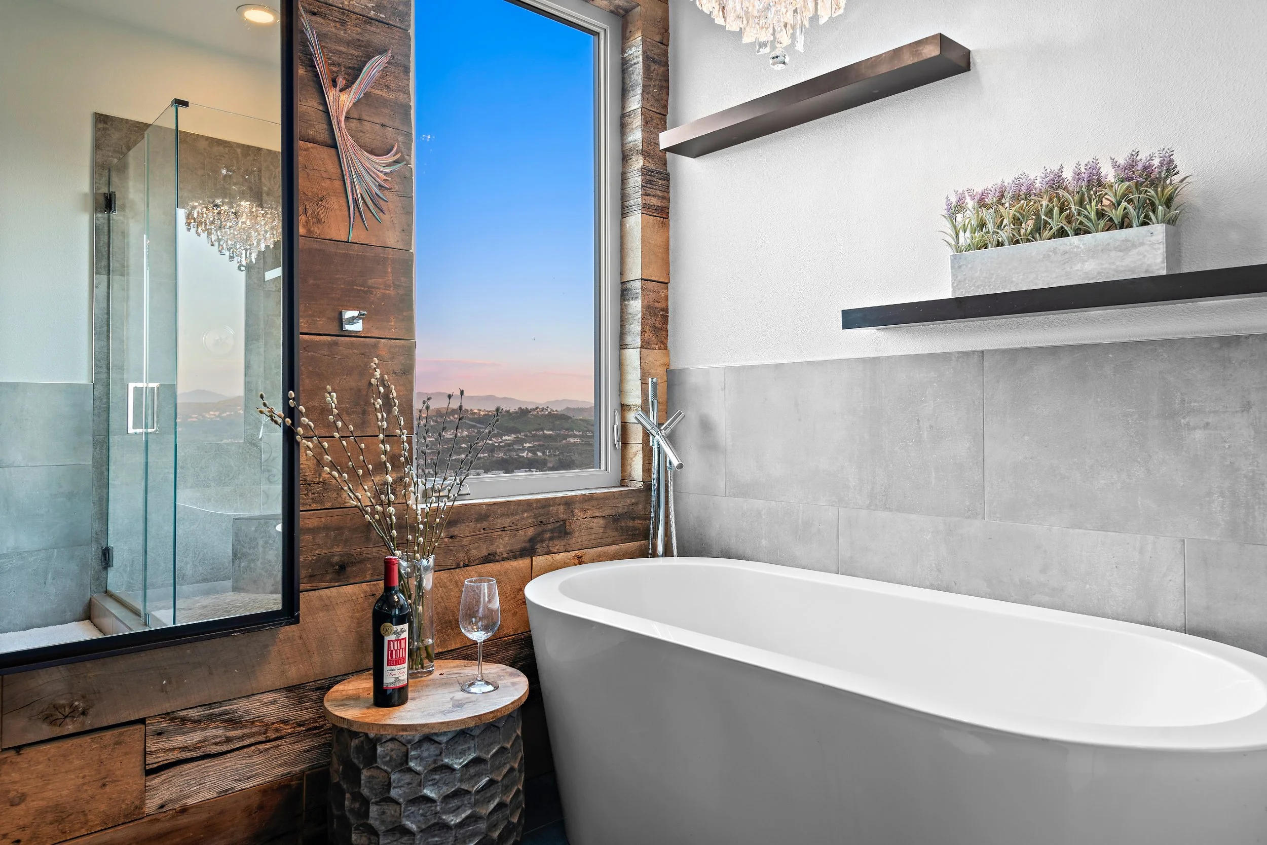 Modern bathroom with a white bathtub, large window showing a sunset view, wooden wall accents, glass-enclosed shower, and decorative shelves with plants.