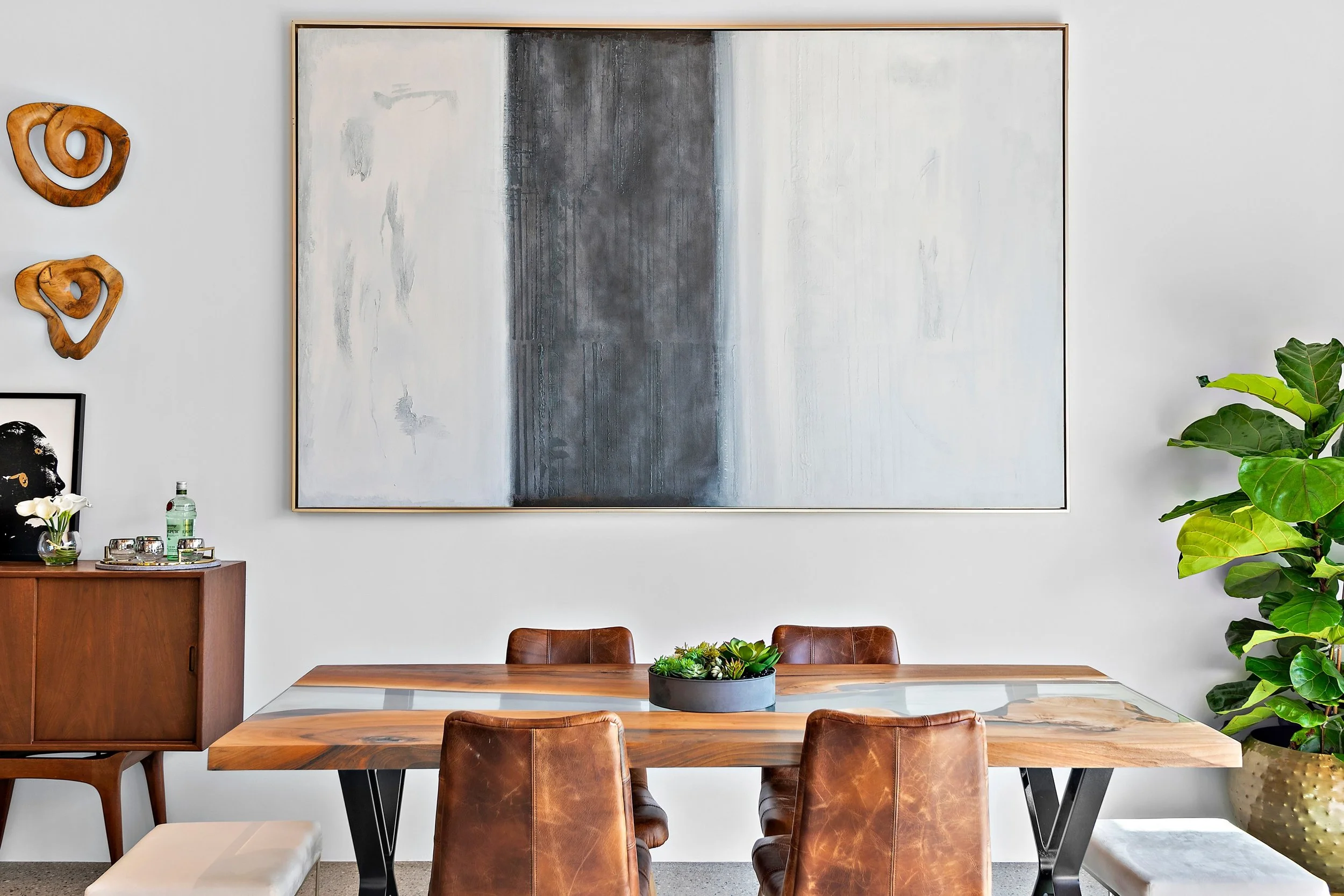 Modern dining room with wooden table, leather chairs, large abstract painting on the wall, wooden wall art, sideboard with glassware and a bottle of water, a framed picture, and a large potted plant.