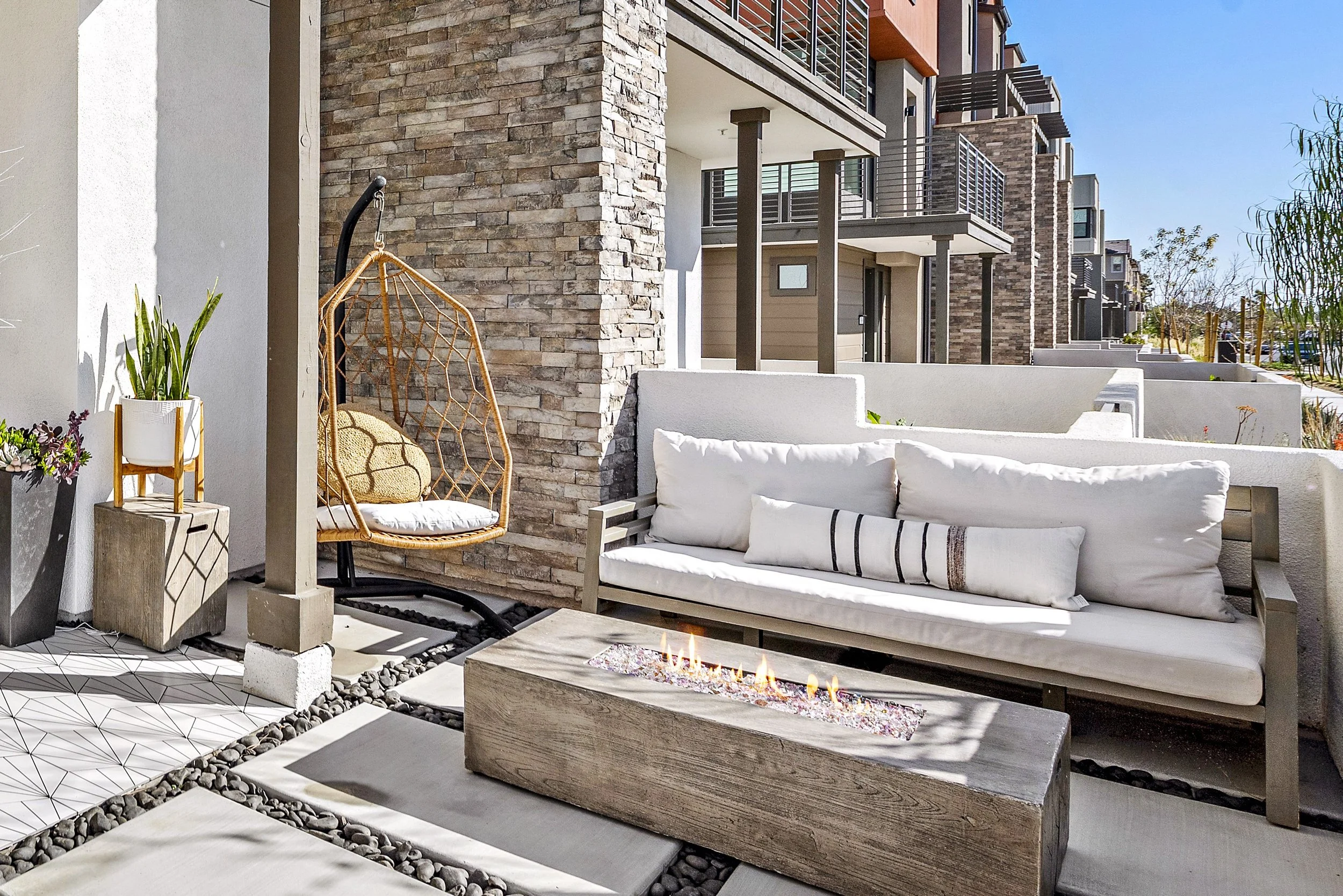 Outdoor patio with a cushioned bench, hanging wicker chair, fire feature, potted plants, and modern residential building in the background.