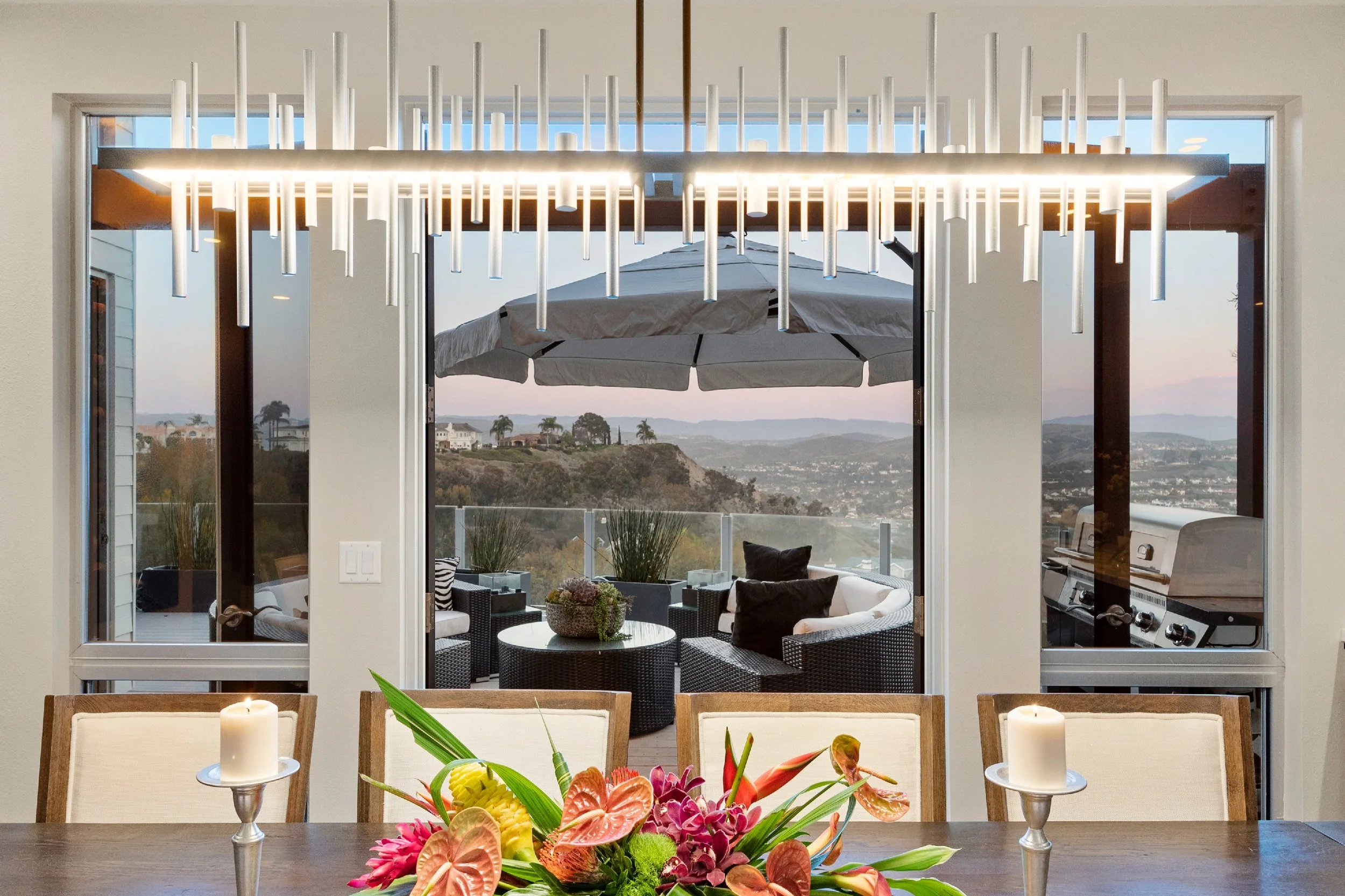 Indoor dining room with a view through large sliding glass doors to an outdoor patio with seating, umbrella, and a scenic landscape of hills and houses in the distance.