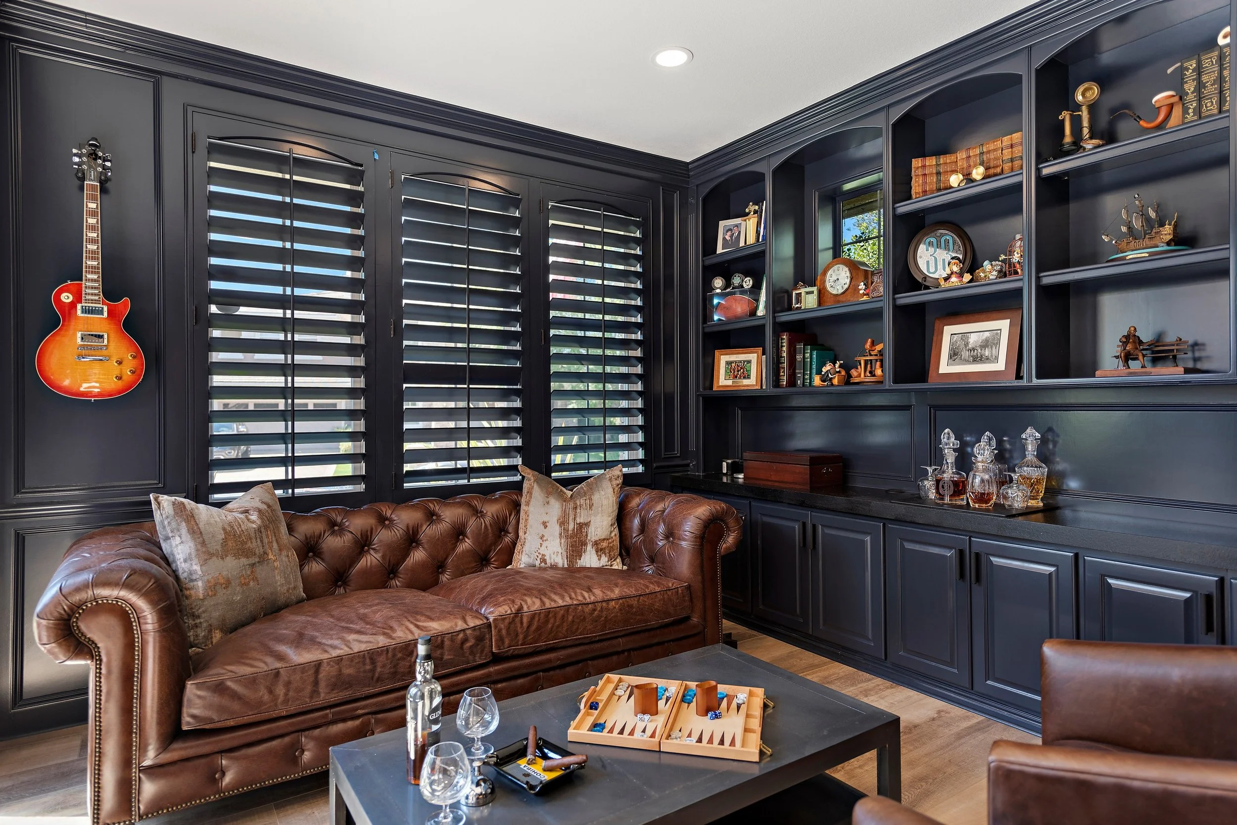 Living room with black paneled walls, large windows with black shutters, a brown leather tufted sofa with pillows, a black coffee table with glass bottles, glasses, a backgammon game, and stored liquor bottles, and a built-in black bookshelf filled w