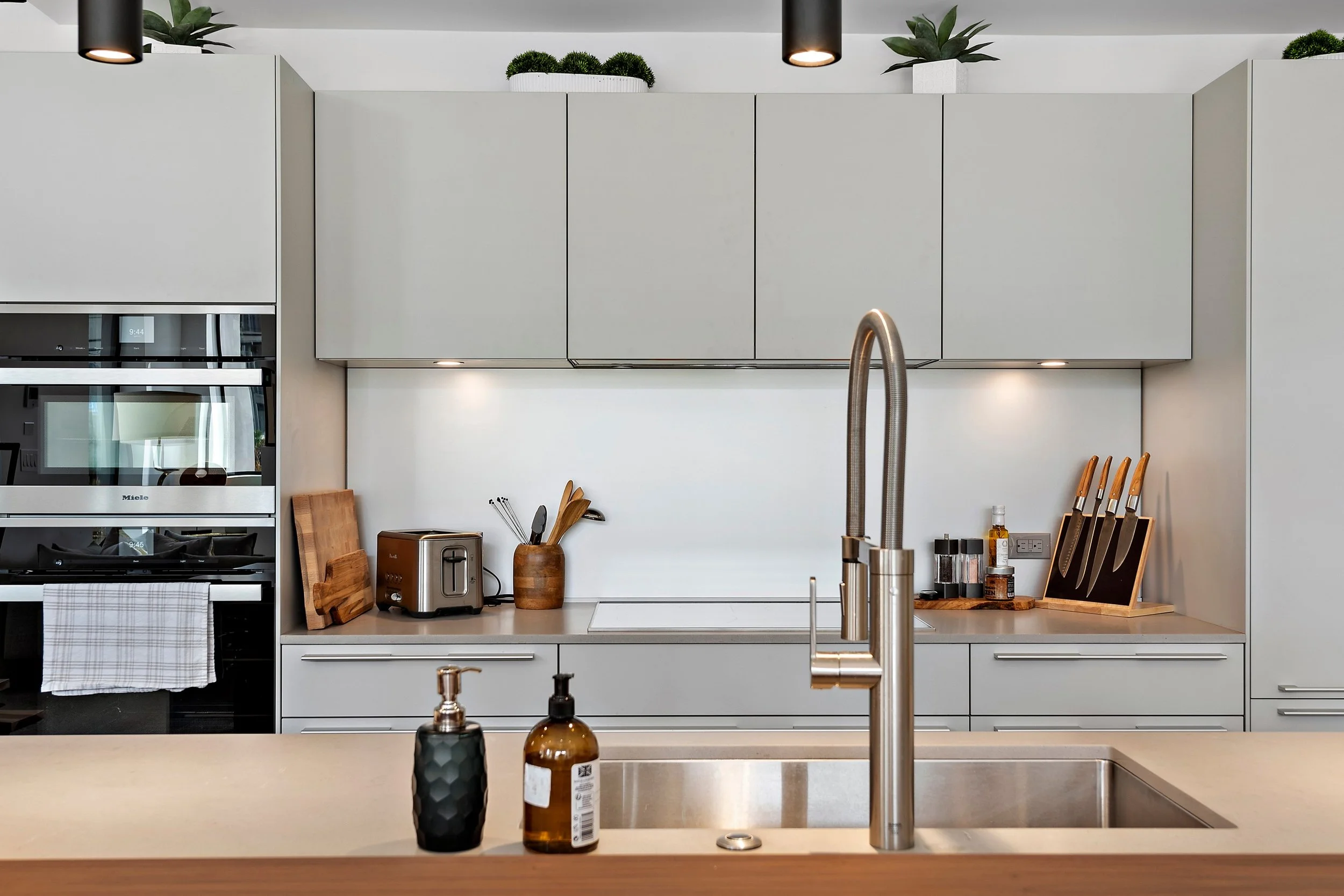 Modern kitchen with white cabinets, stainless steel sink, black faucet, and various kitchen appliances including a toaster, knife set, and bottles, with decorative plants on the top cabinets.