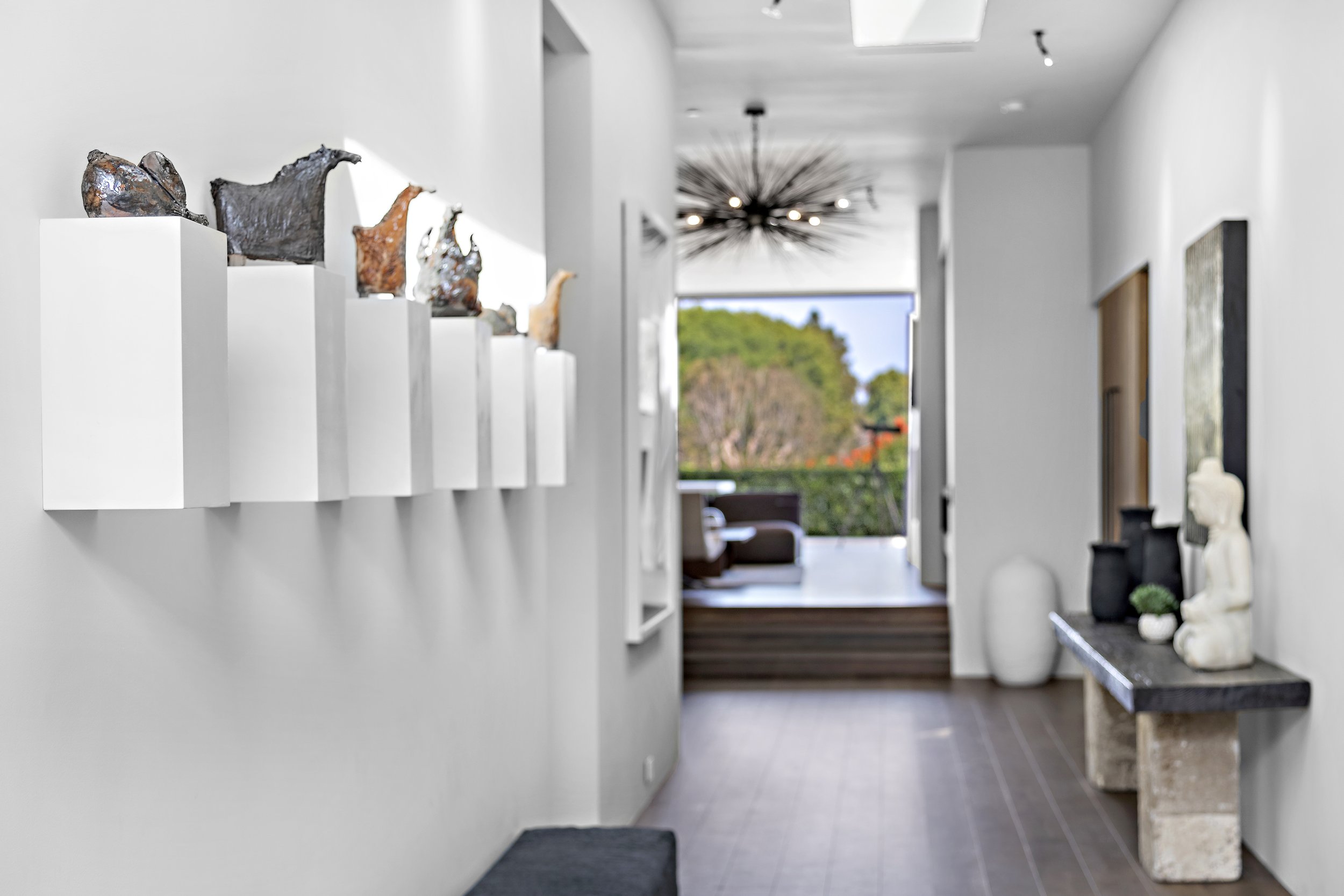 Interior of a modern, minimalist home with white walls, decorative sculptures, and a view of a patio with greenery outside.