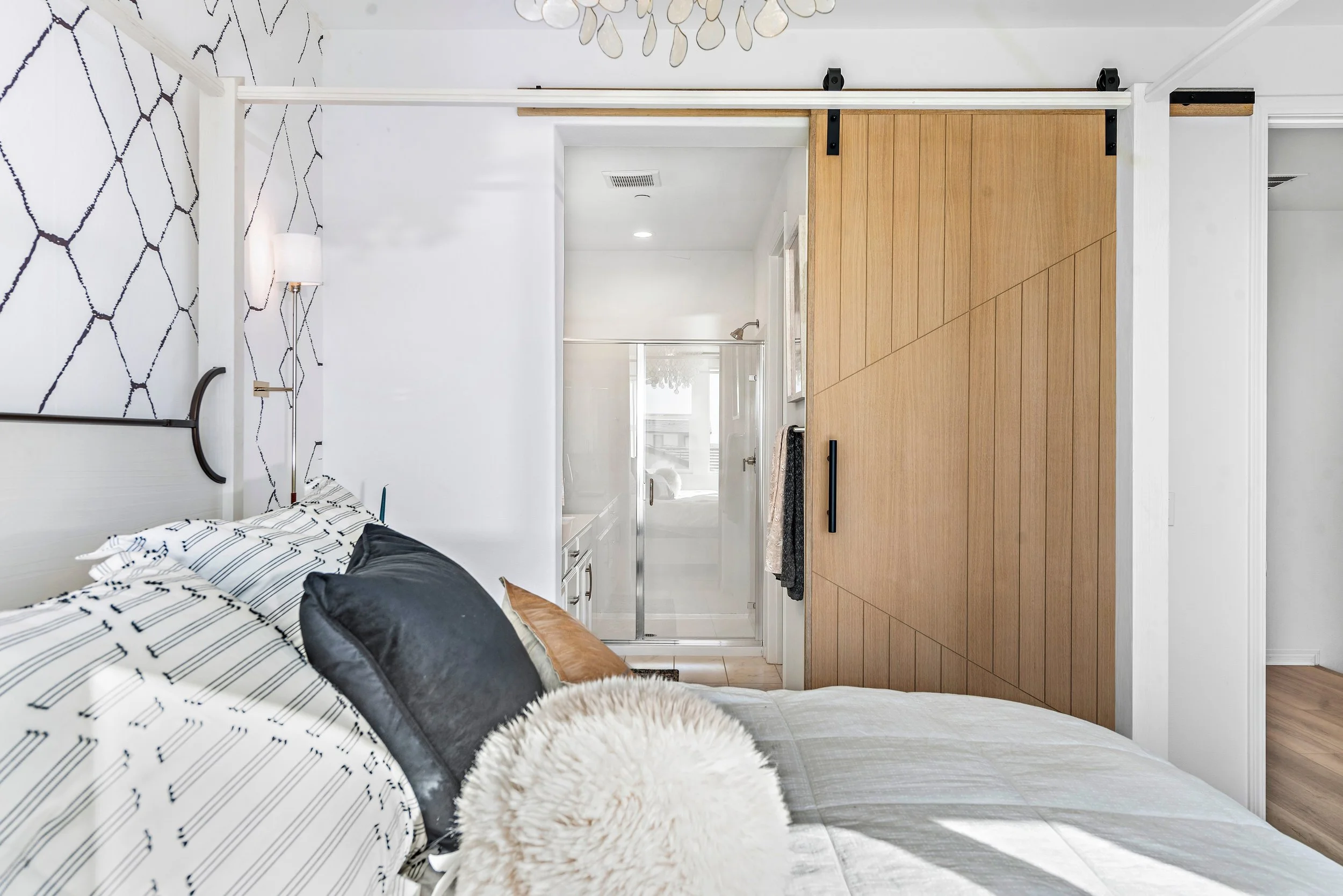 A bedroom with a partially open sliding wooden barn door leading to a bathroom with a walk-in shower and a window. The bed has black, white, and beige bedding, and there are wall-mounted lamps and patterned wallpaper on the left wall.