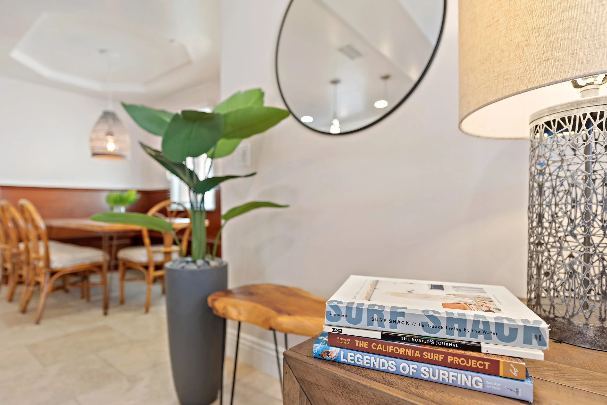 Stack of four books on a wooden table next to a ceramic table lamp with a beige shade. A large leafy plant in a gray pot is beside the table. In the background, a dining area with wooden chairs and a dining table is visible, along with hanging pendan