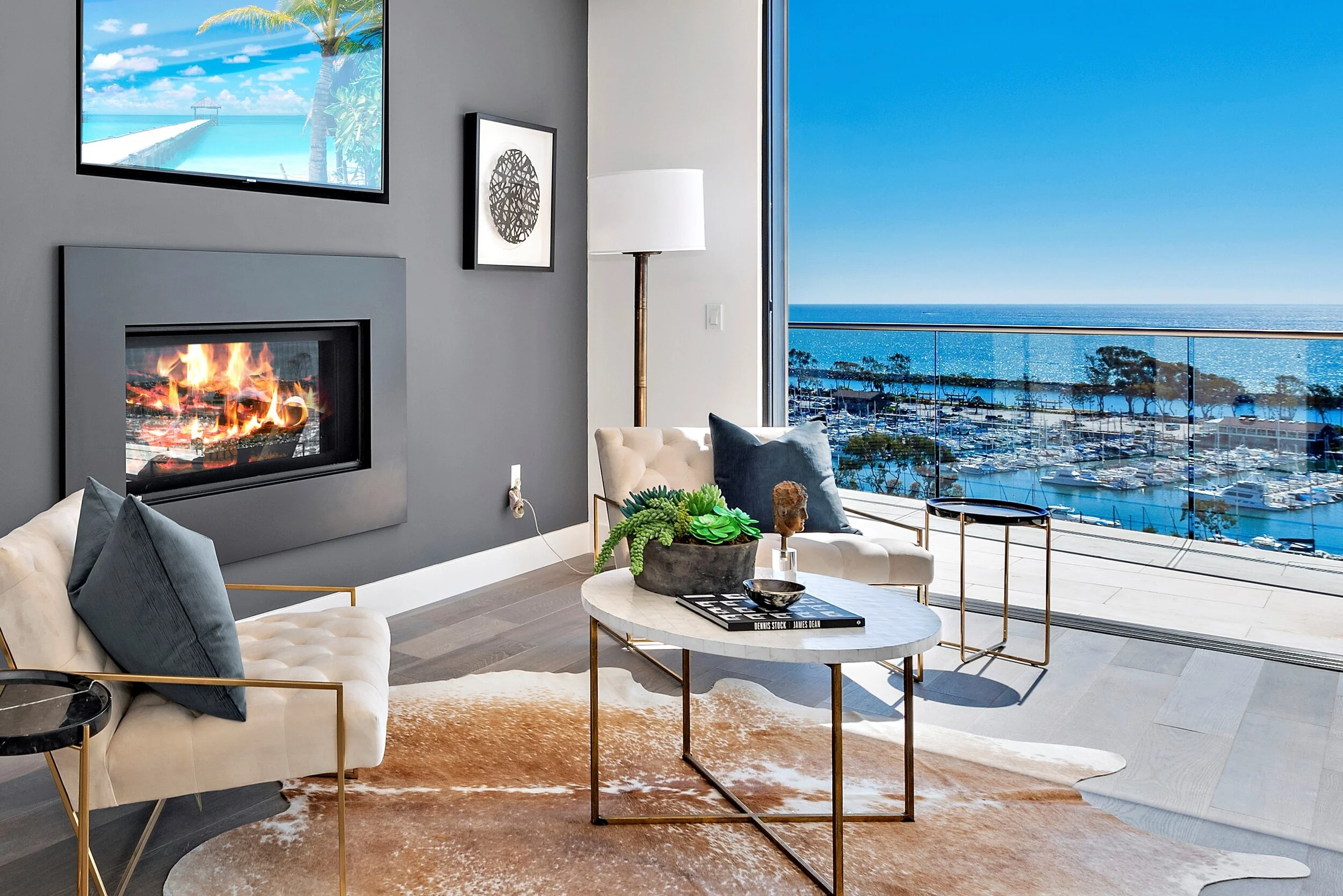 Living room with modern decor overlooking a marina and ocean through a glass balcony door, featuring a gas fireplace, two white sofas with dark cushions, a round marble coffee table with books and a plant, a tall floor lamp, and a large wall-mounted 