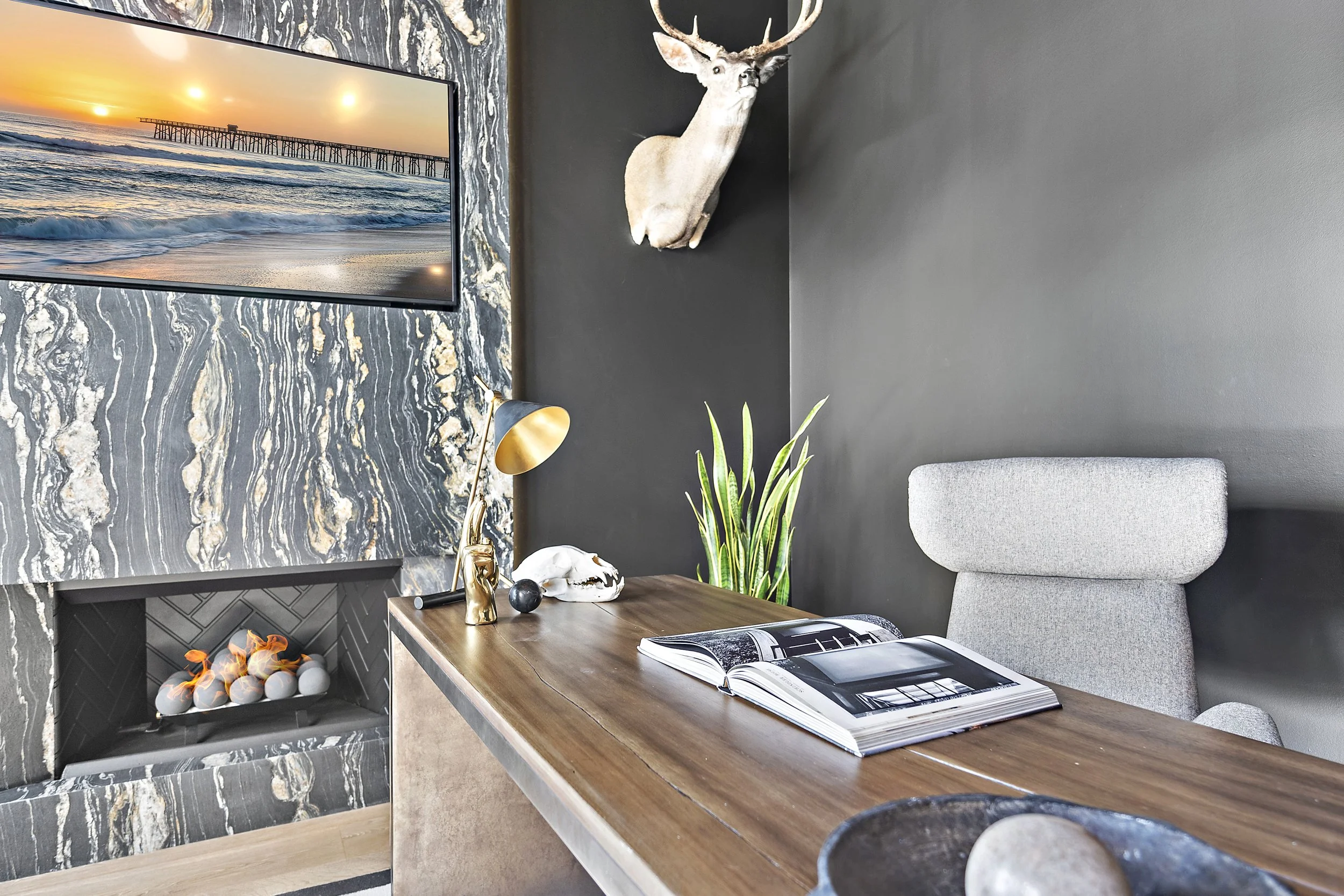 Modern home office with a wooden desk, open magazine, decorative skull, gold desk lamp, potted plant, and a mounted deer head on a dark gray wall. A large flat-screen TV displays a sunset over a pier and ocean, and a fireplace with firewood is below 