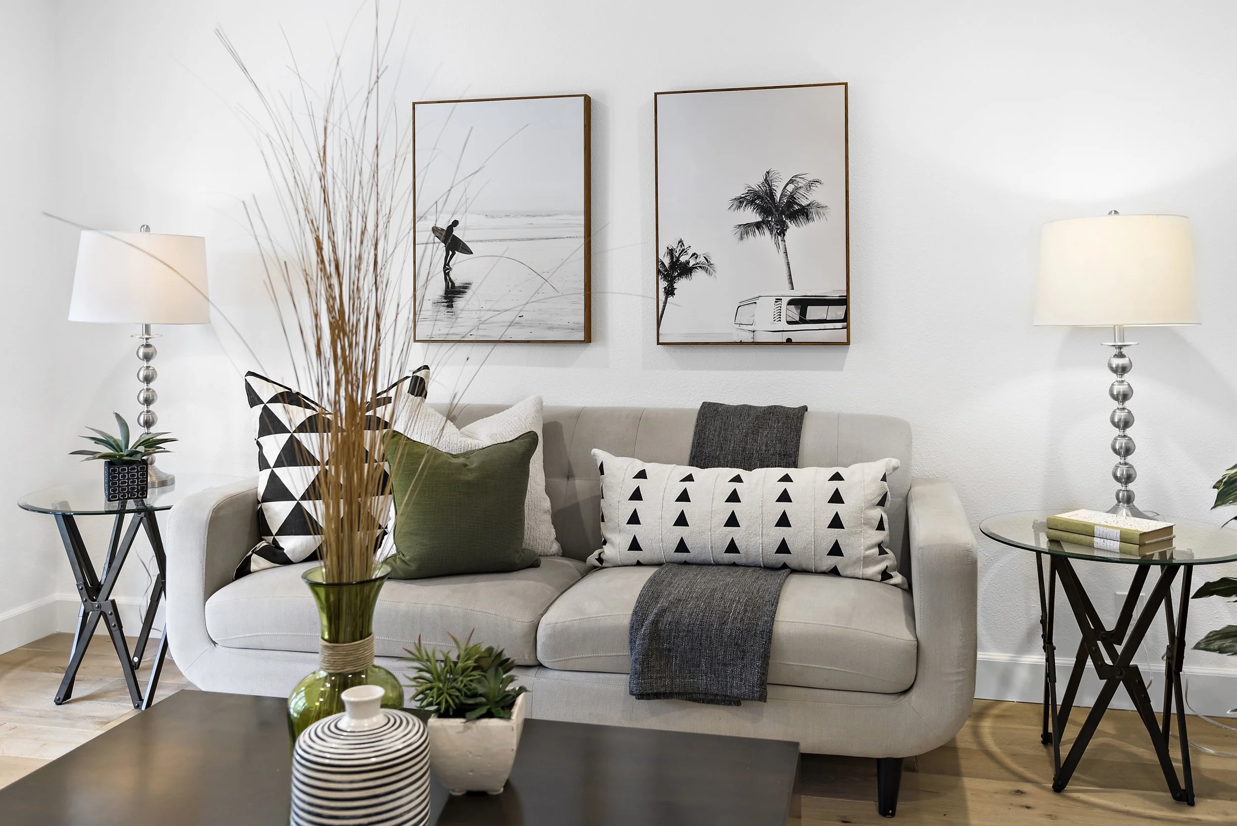 Living room with a light-colored sofa, decorative pillows, plants on the coffee table, and framed black-and-white photos of a surfer and palm trees on the wall.