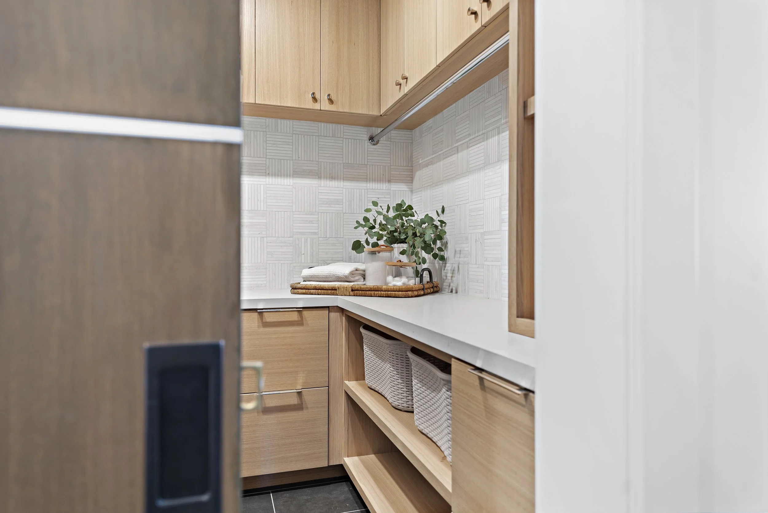 Part of a kitchen with wooden cabinetry, a white countertop, decorative backsplash, green plant, and storage baskets.
