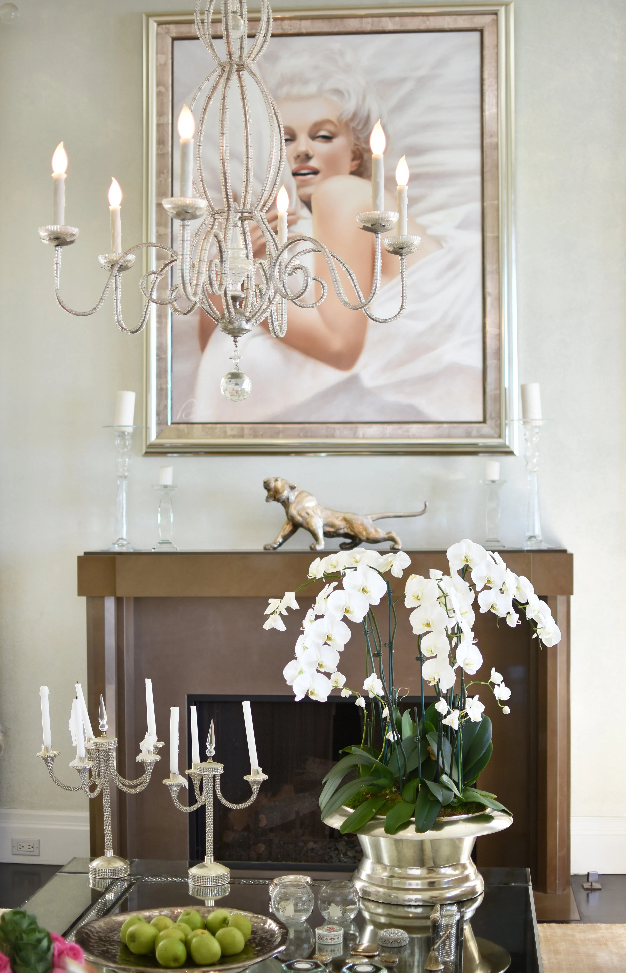 Living room interior with a chandelier, a large portrait of Marilyn Monroe above the fireplace, a decorative sculpture of a dog on the mantle, and a white orchid plant in a silver pot on the coffee table. The photo also shows a silver candelabrum wit