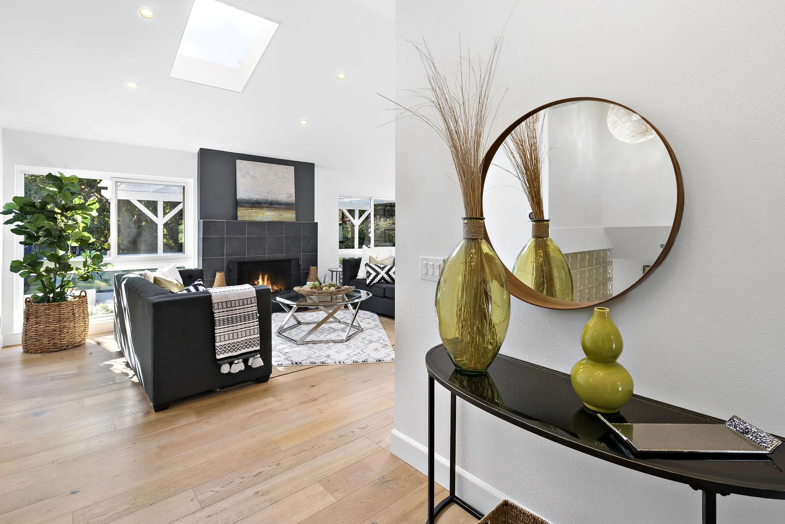 Living room with a fireplace, black sofas, large windows, and a potted plant, with a decorative table and artwork above the fireplace. In the foreground, a black console table with three yellow vases and a round mirror above.