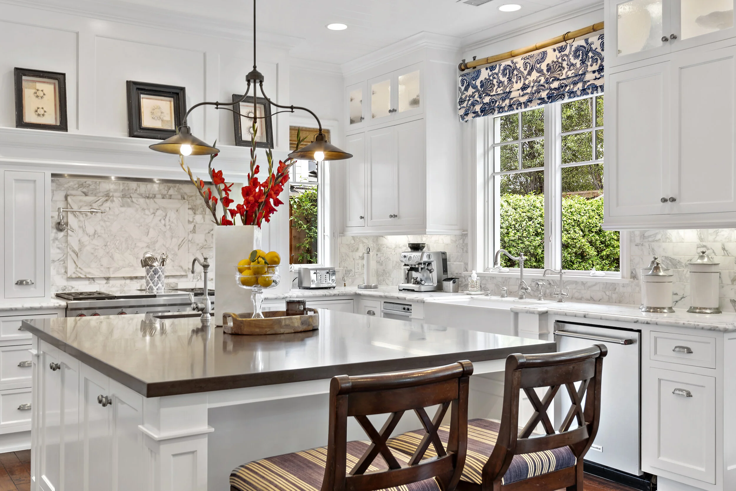 Bright white kitchen with large window, white cabinets, marble backsplash, and wooden island counter. Decor includes red flowers in a white vase, framed pictures, and blue and white window valance.