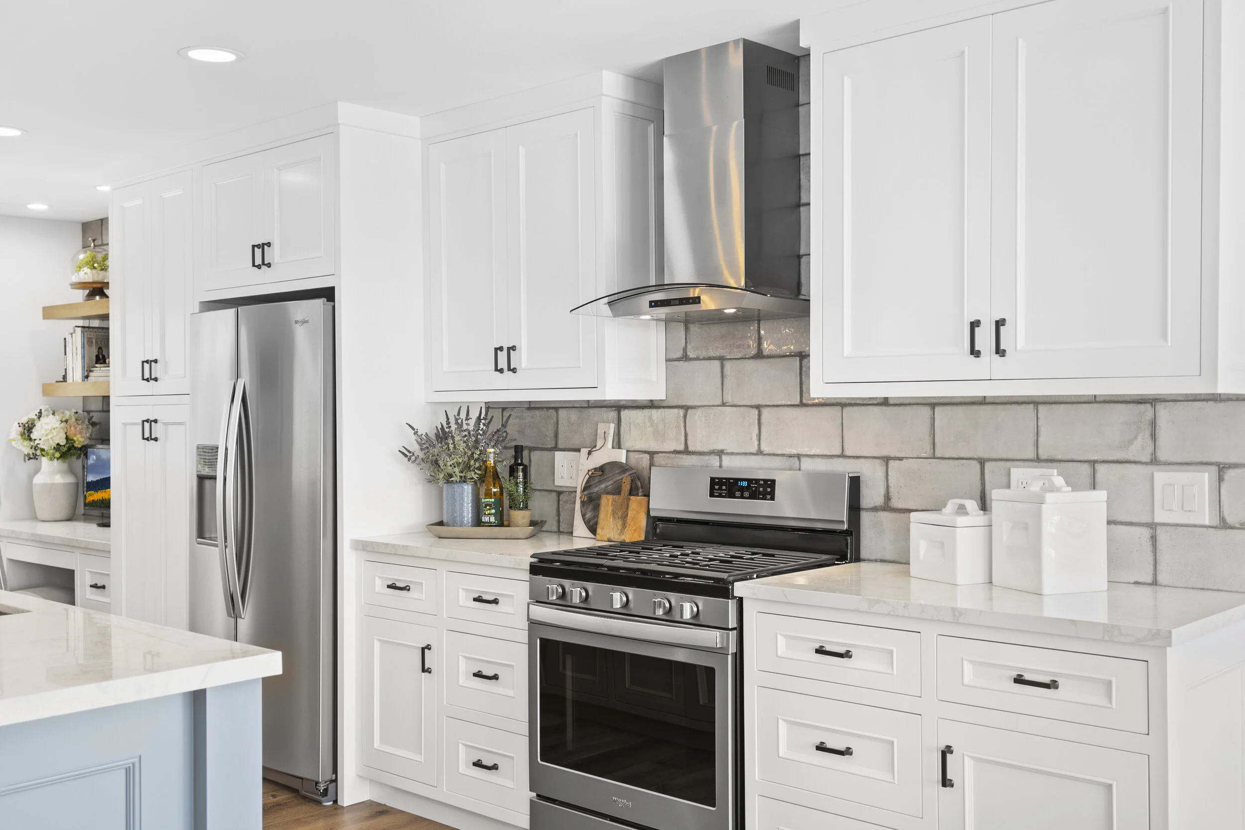 Modern kitchen with white cabinets, stainless steel refrigerator, stove, and hood, gray brick wall backsplash, and decorative items on the countertop.