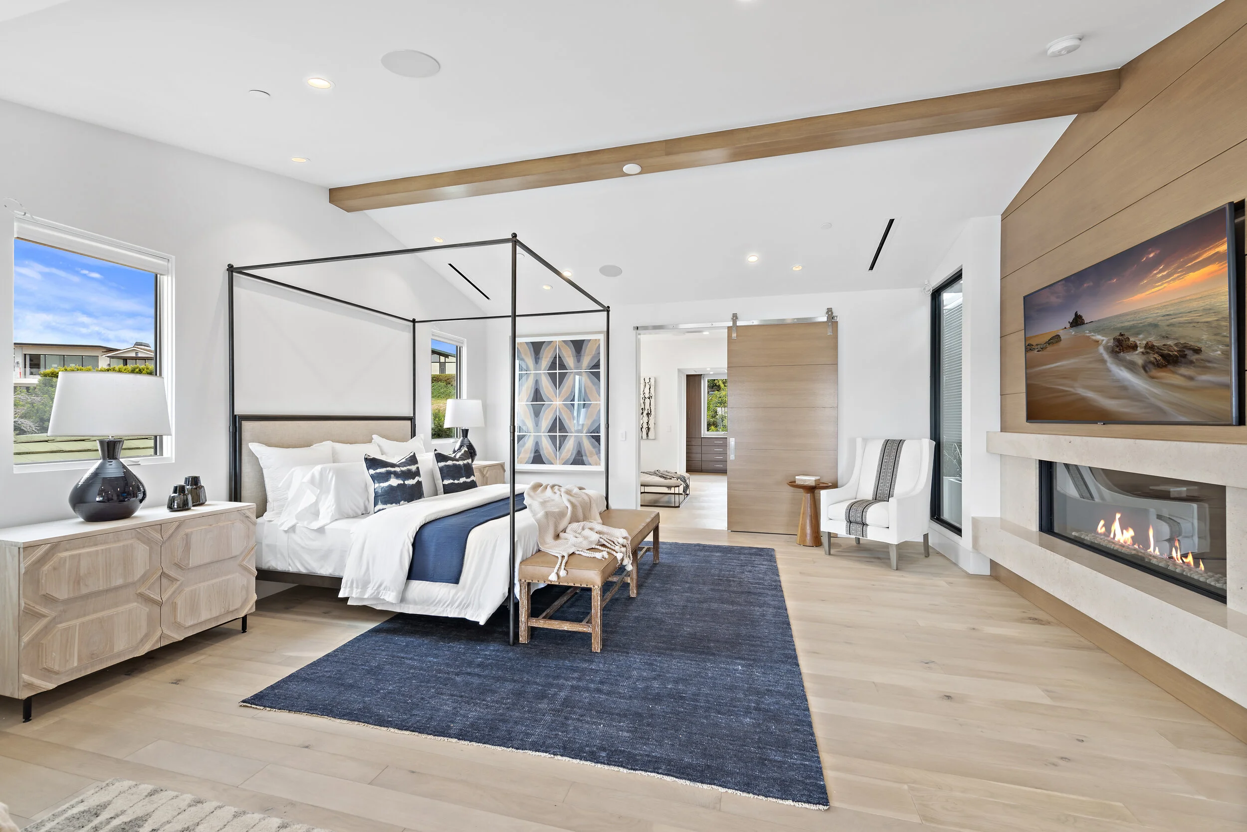 Modern bedroom with a canopy bed, blue and white color scheme, a fireplace, a wall-mounted TV showing a beach scene, and large windows letting in natural light.