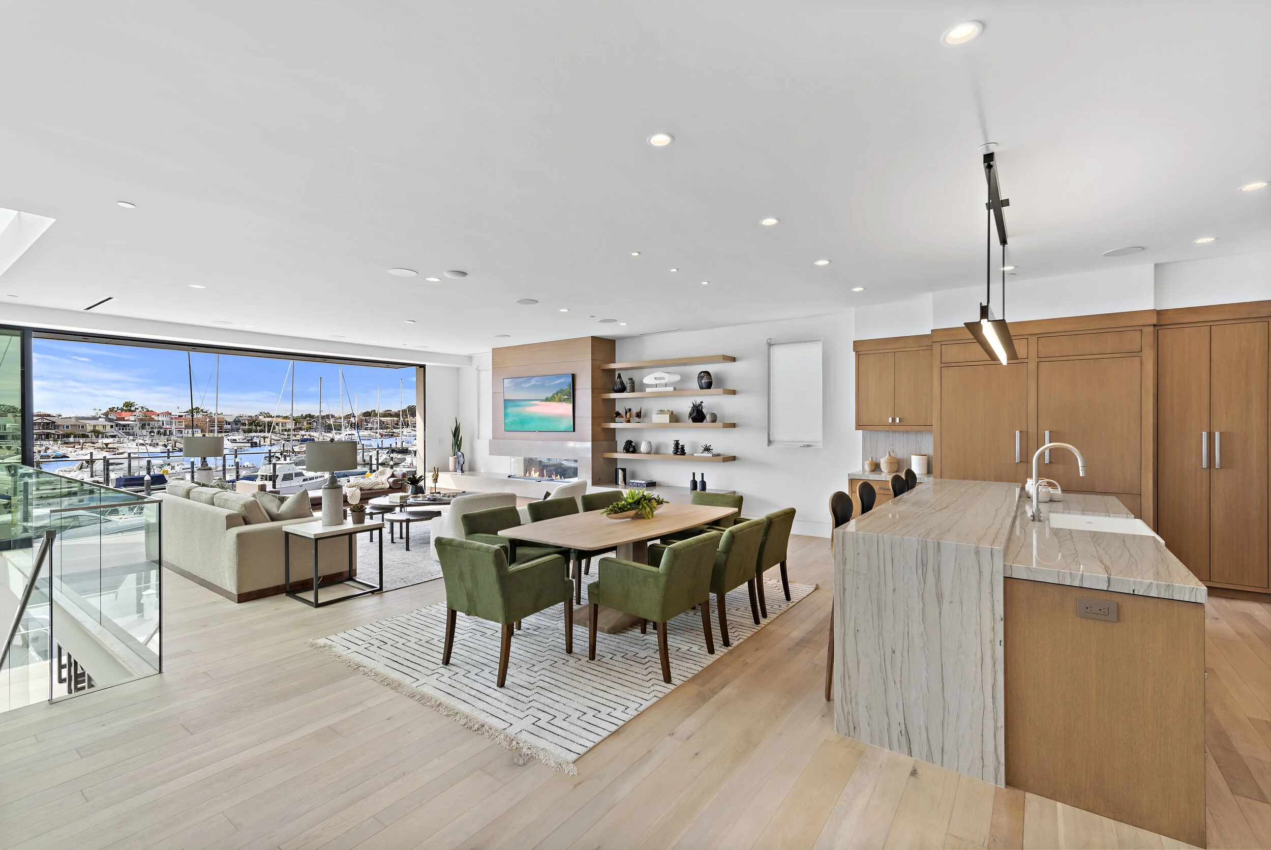 Open-concept living and dining area with large window view of marina, modern fireplace, mounted TV, shelves with decor, green chairs, beige sofa, kitchen with marble island and wooden cabinets.