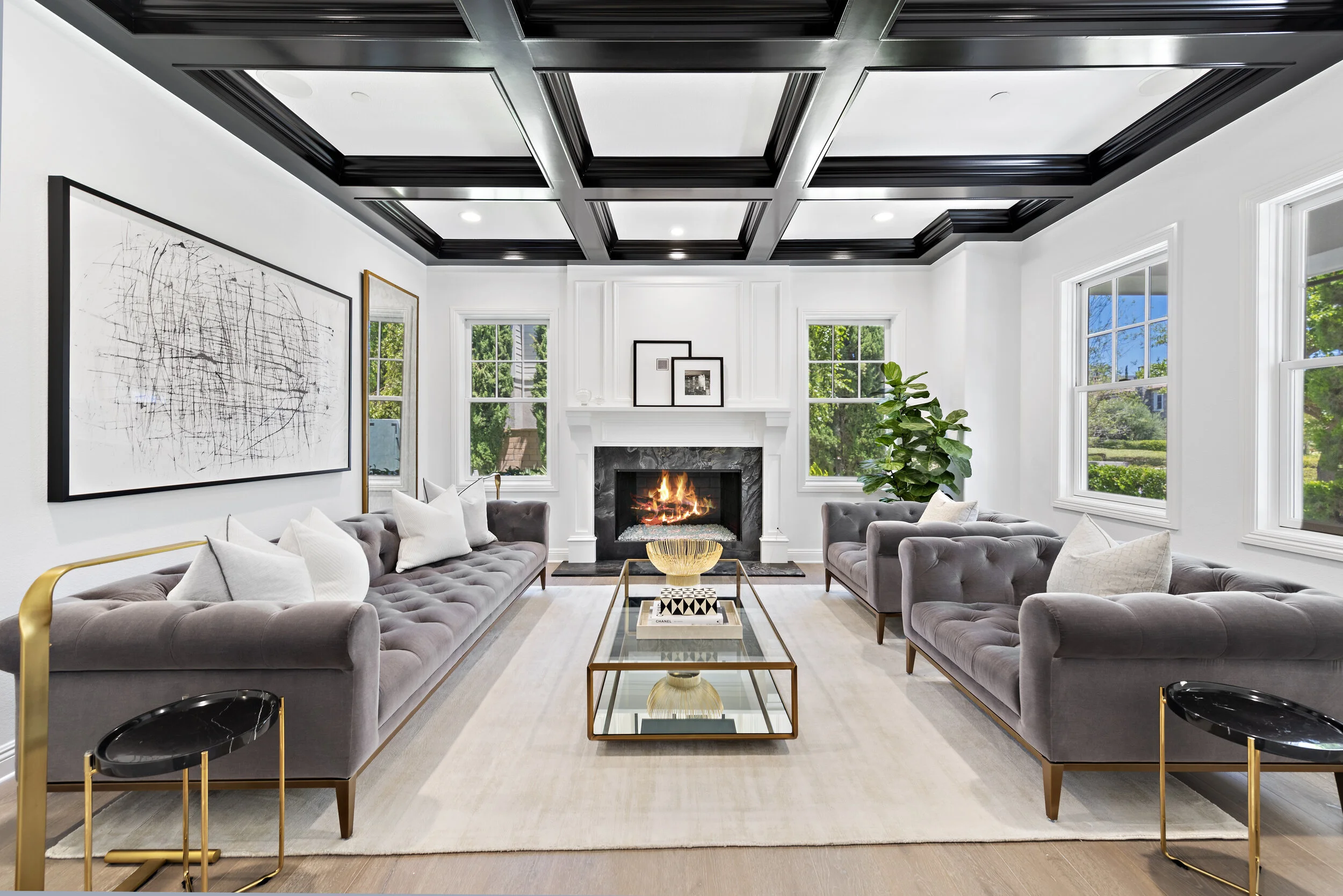 Modern living room with four gray velvet tufted sofas, black and white abstract art, a white fireplace with a fire, three windows showing green trees, a glass coffee table with decor, beige rug, black and gold side tables, a large green plant, and bl