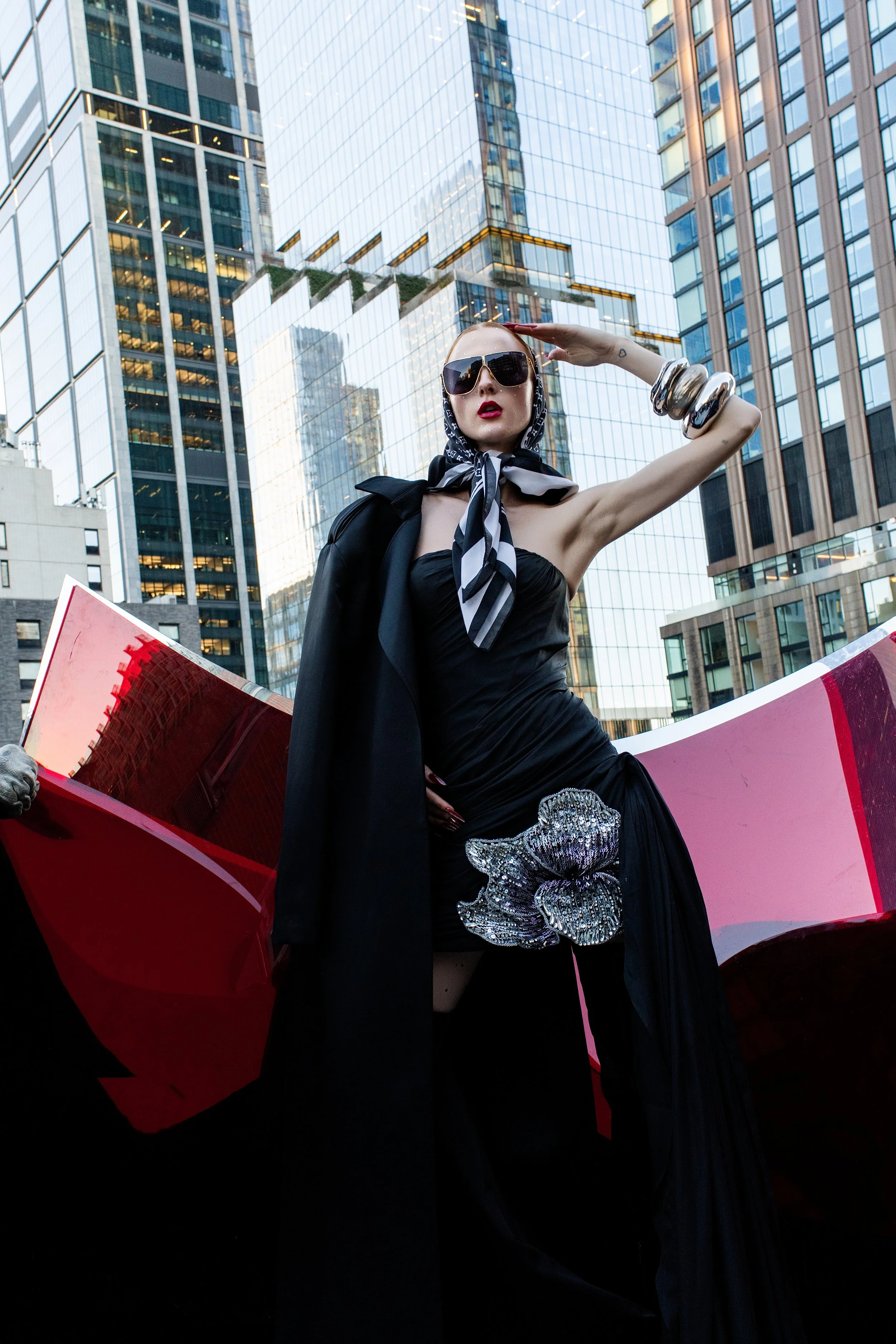 Una mujer elegante con gafas oscuras y bufanda de rayas en un entorno urbano con edificios de cristal.