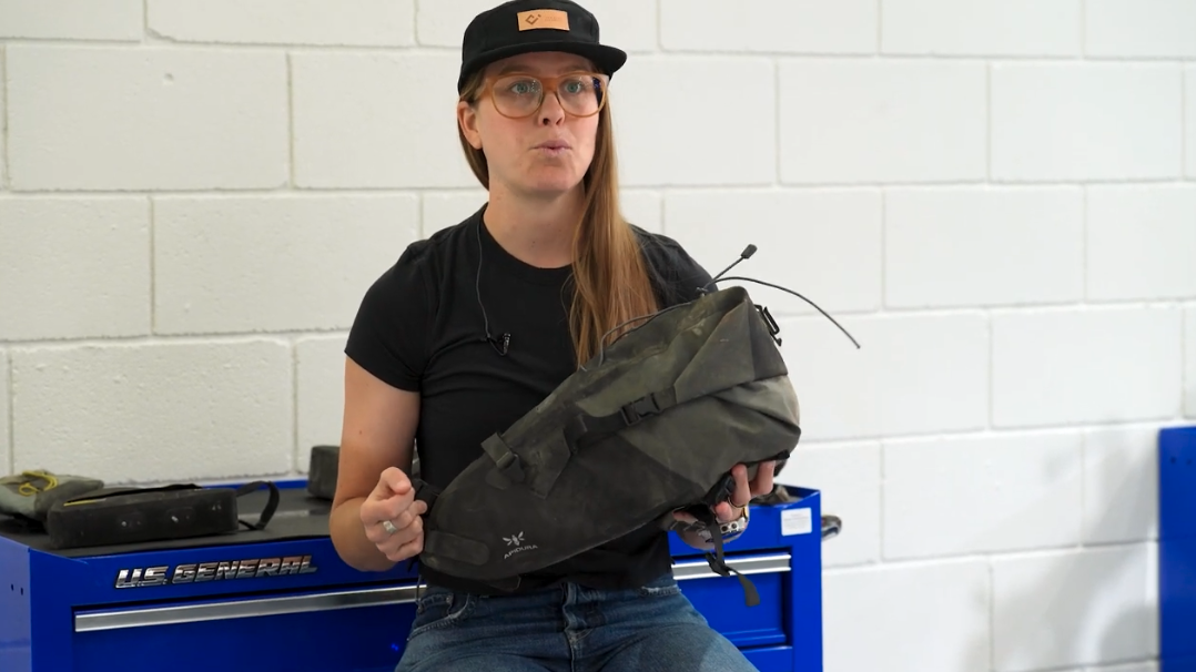 A woman wearing a black cap, glasses, and black t-shirt holds up an olive green bikepacking saddle bag while speaking, seated in front of a blue U.S. General tool chest against a white cinder block wall.