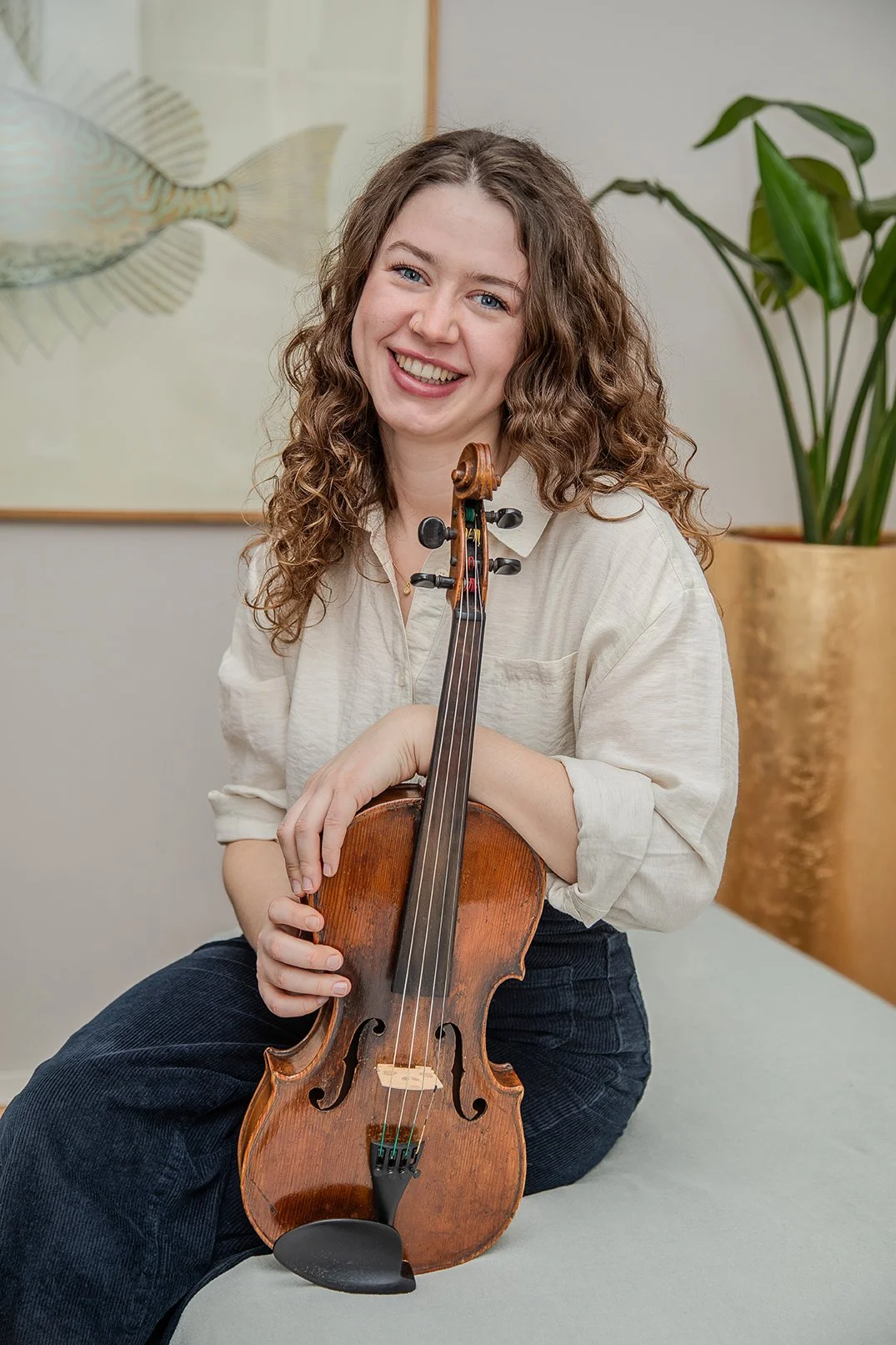 Eine lachende junge Frau mit lockigem Haar hält eine Bratsche vor sich, sitzt auf einer Bank in einem Raum mit Pflanzen und einem Wandbild.