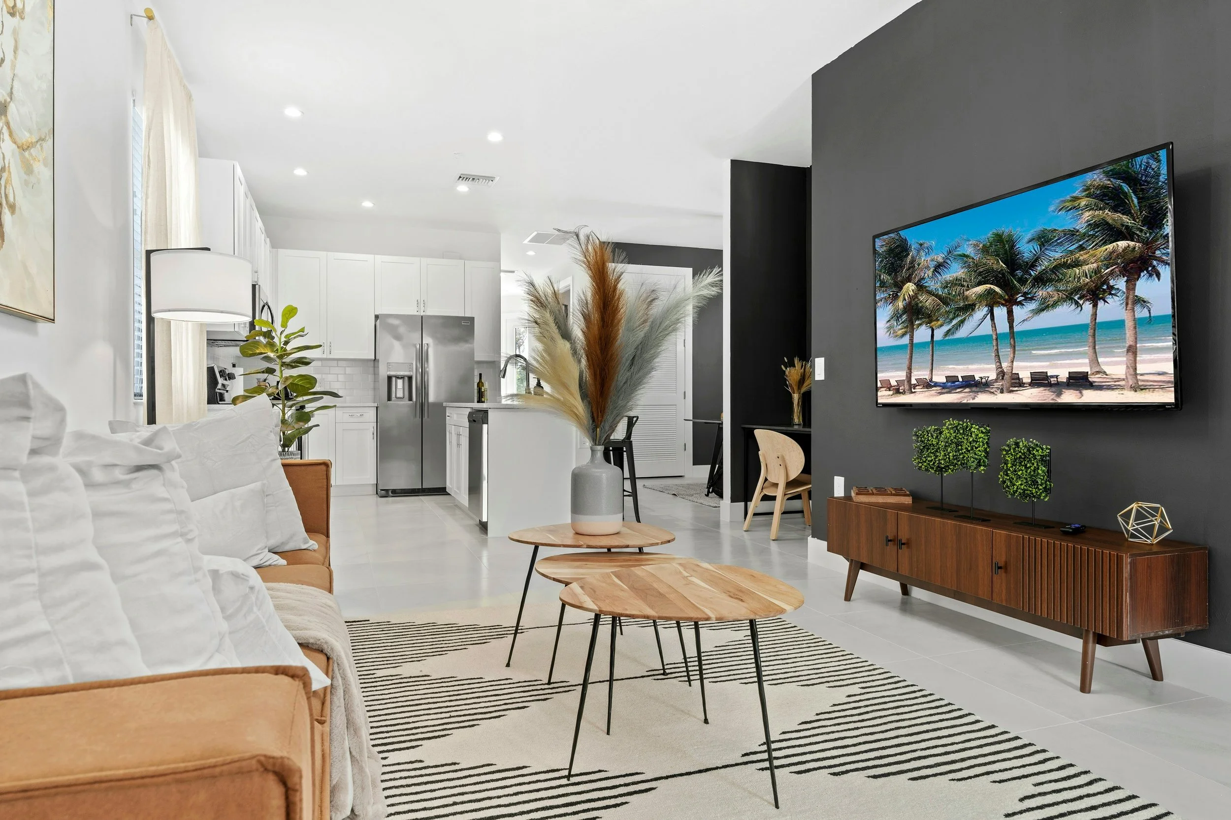 Living room with beige sofa, wooden coffee tables, black and white striped rug, TV on a dark wall showing beach scene, kitchen with white cabinets and stainless steel appliances, decorative plants and pampas grass in a vase.