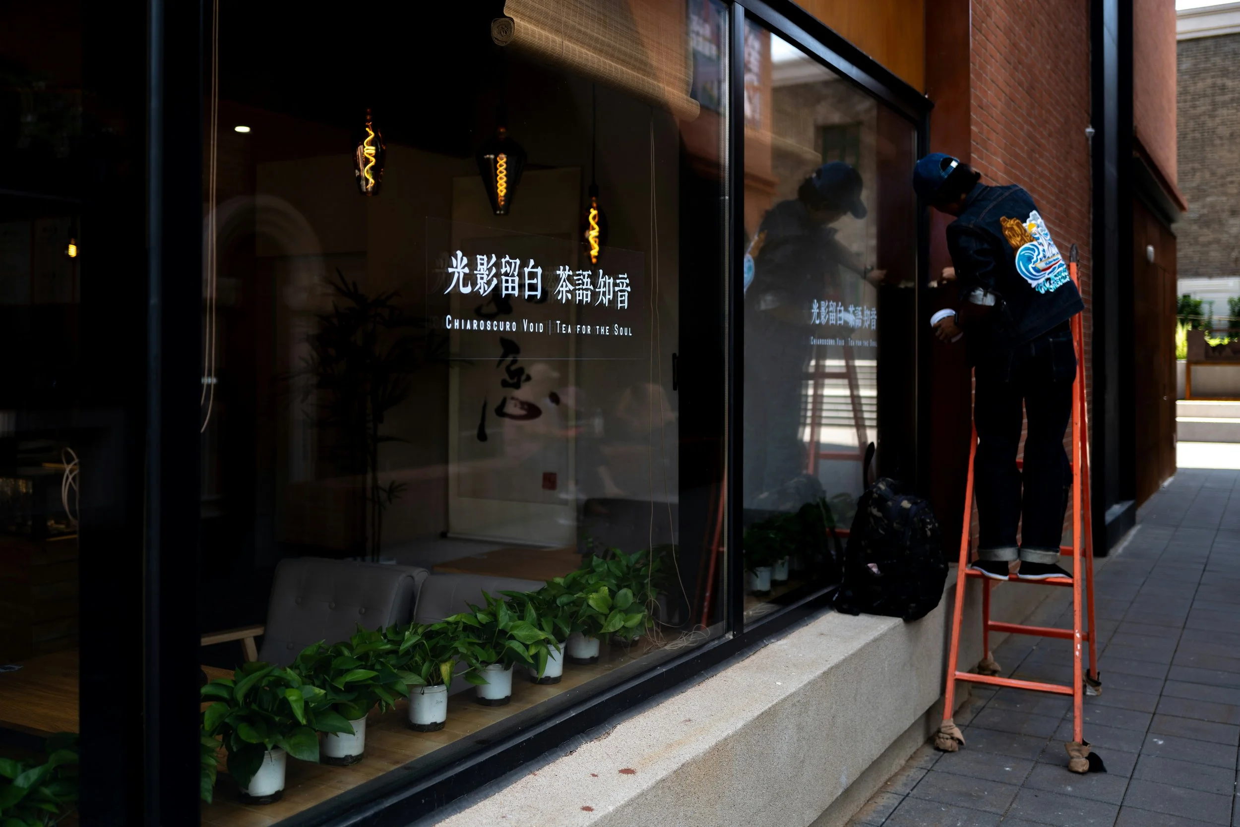 A person standing on a ladder outside of a shop window, possibly working on signage or decoration, with the shop's name in both Chinese characters and English reading 'Chiaroscuro Void | Tea for the Soul.' The shop interior has modern lighting and indoor plants are visible.