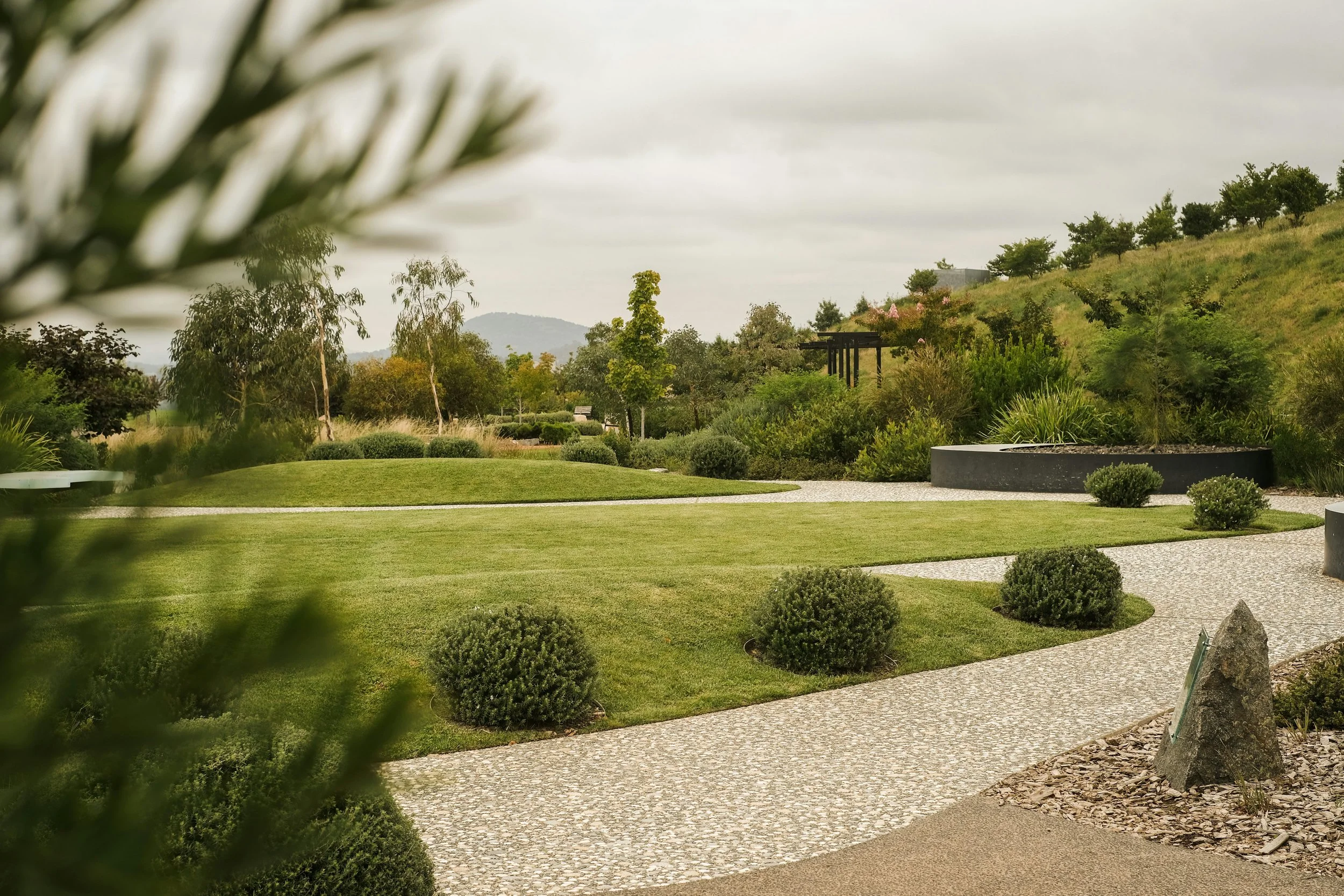 A landscaped garden with a curved pebble pathway, small bushes, trees, and a grassy hill in the background, under an overcast sky.