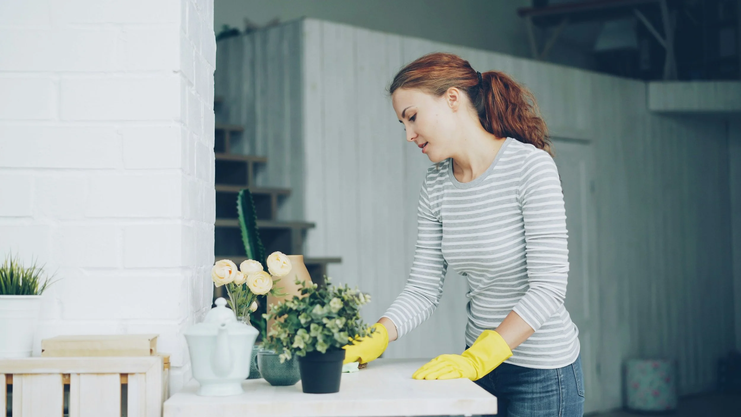 Woman arranging potted plants on a white kitchen countertop, wearing yellow gloves and a striped shirt.