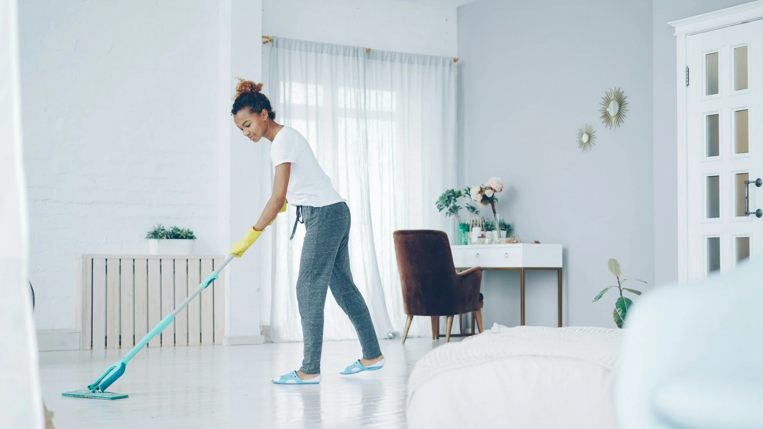 A woman wearing yellow rubber gloves and slippers is mopping a white floor in a bright, modern living room with white walls and light-colored curtains.