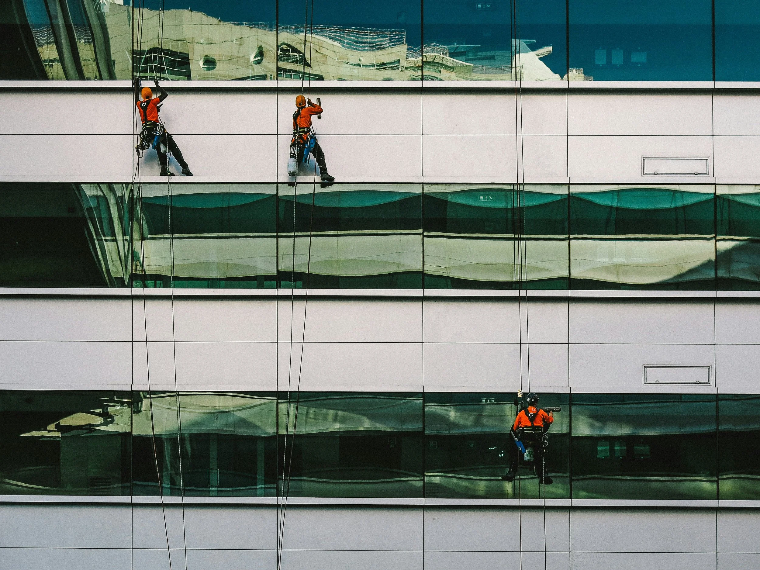 Three window washers cleaning the glass exterior of a modern building with reflective windows.