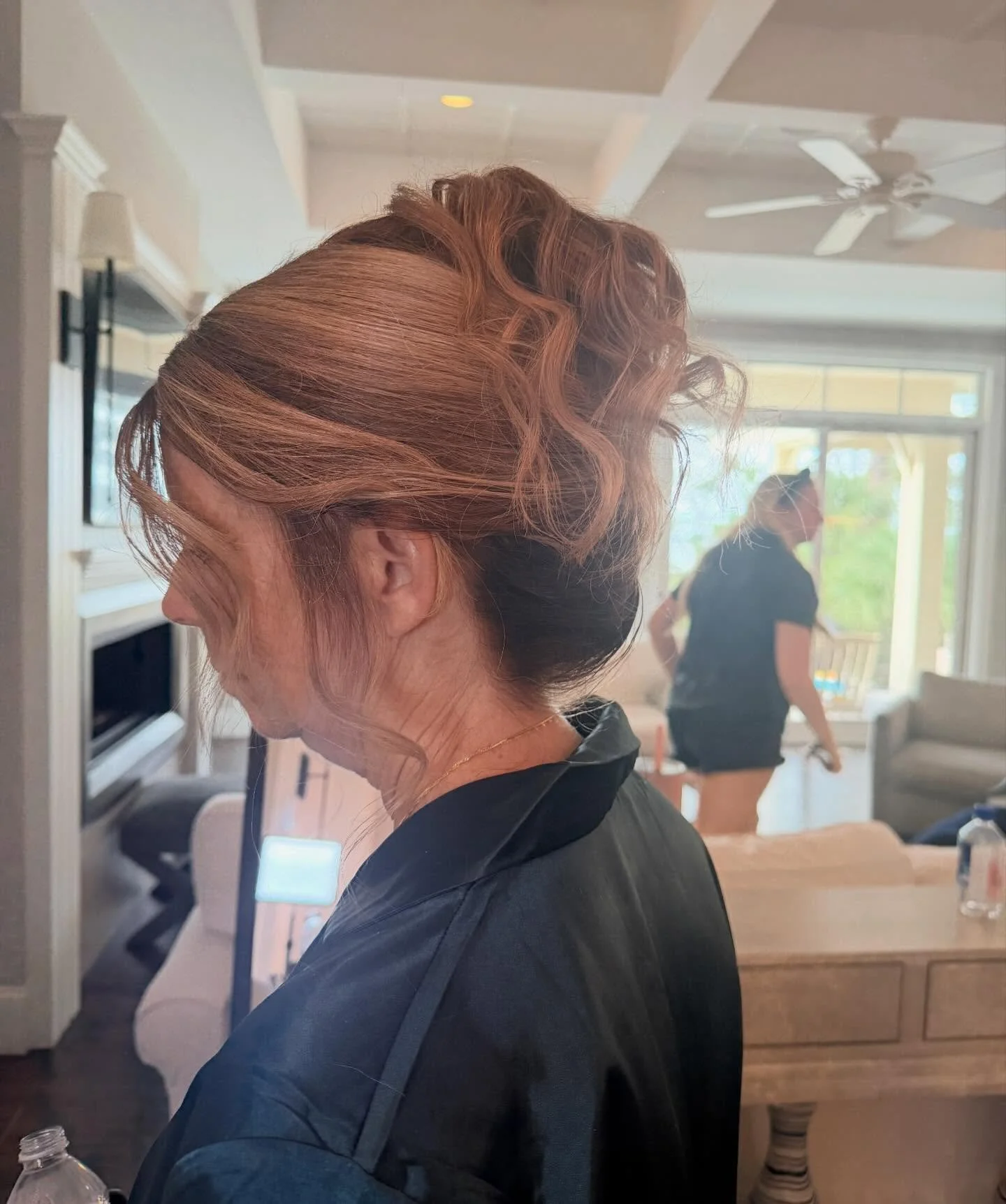 Woman with styled hair in an elegant updo, wearing a dark top, in a bright indoor room with another woman in the background near a window.