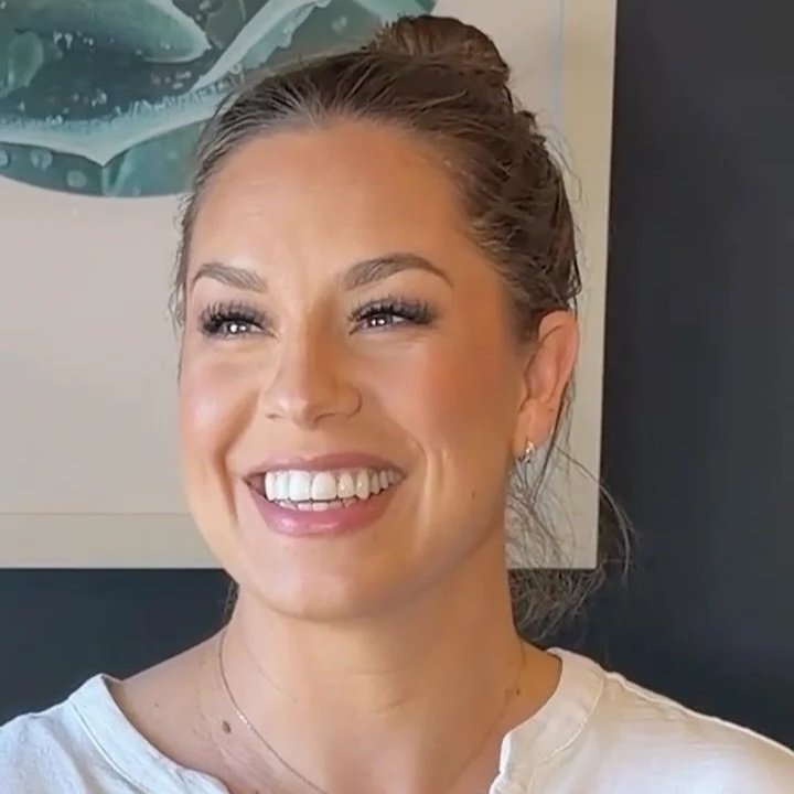 A smiling woman with light skin and her hair tied up, wearing a white shirt and earrings, standing indoors with a piece of artwork in the background.
