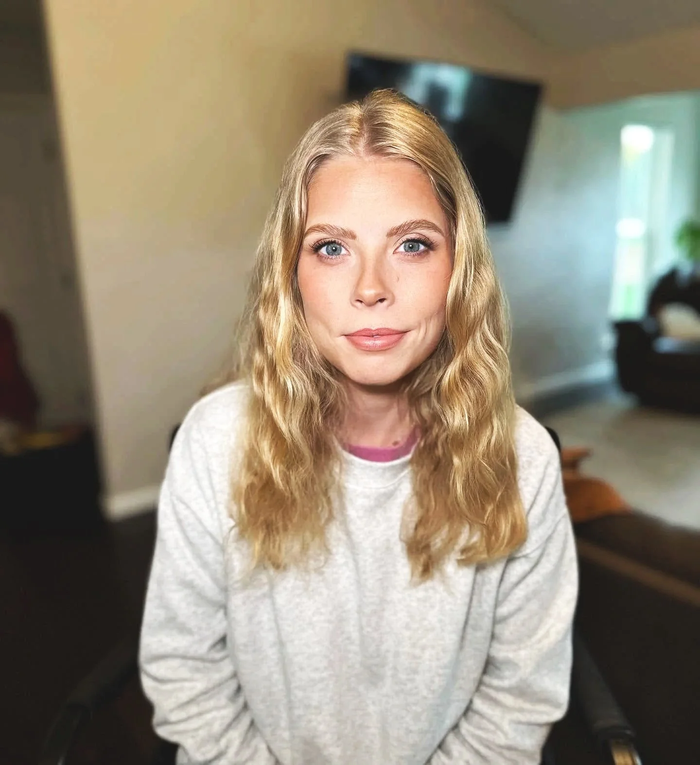 A woman with long, wavy blonde hair and blue eyes sitting indoors with a blurred TV and window in the background.