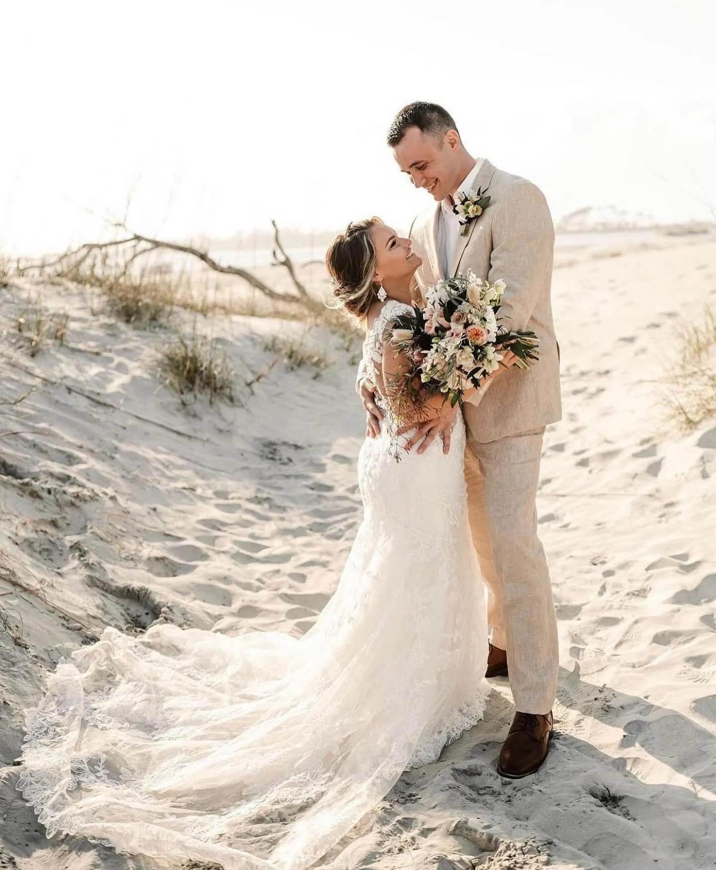 A bride and groom standing on a sandy beach, embracing and smiling at each other. The bride is holding a bouquet and wearing a lace wedding dress, while the groom is dressed in a light-colored suit. The background shows sand dunes and sparse vegetati