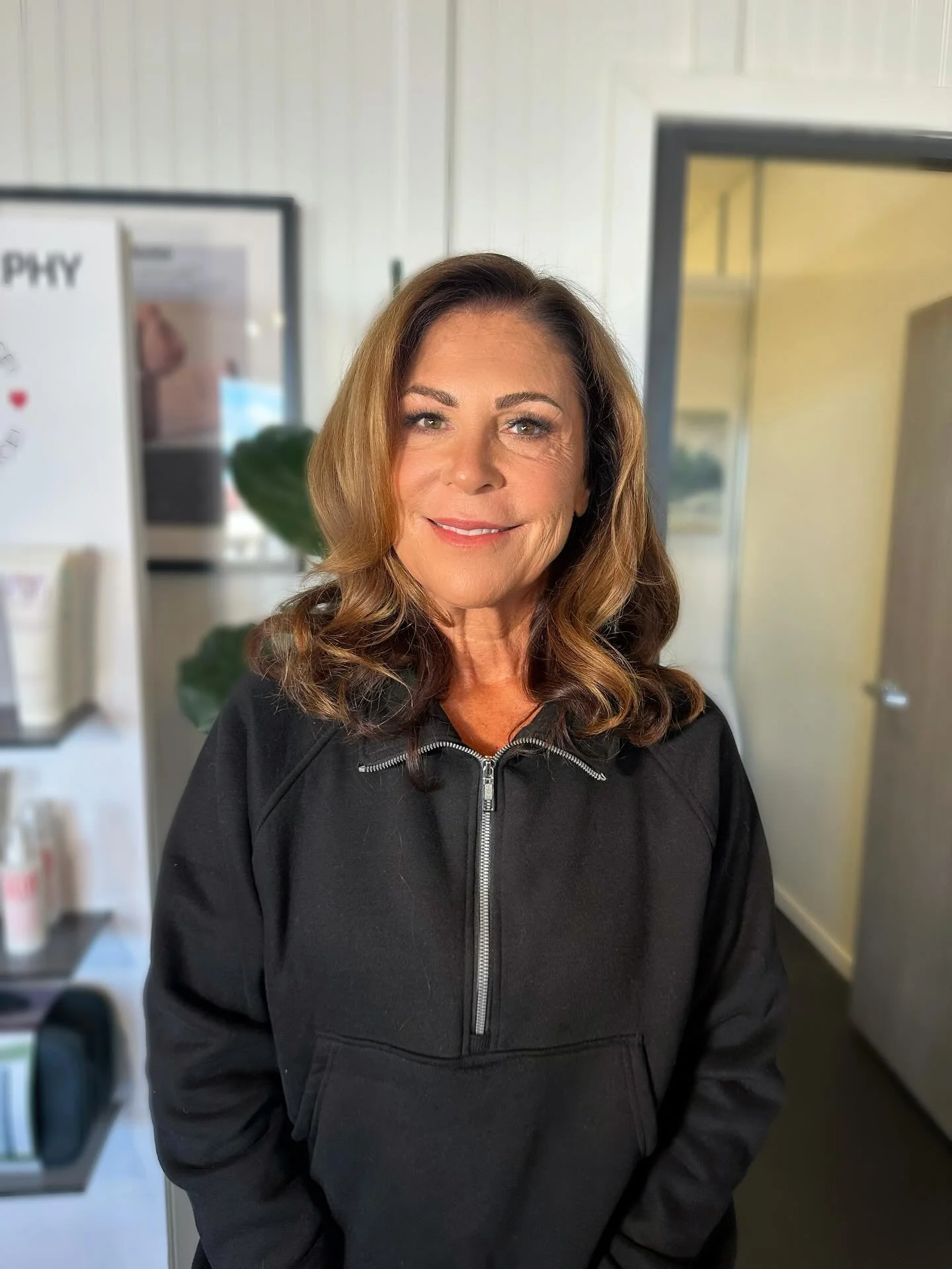 A woman with shoulder-length brown hair and a friendly smile, wearing a black zip-up hoodie, standing indoors in front of a door and a wall with a poster.