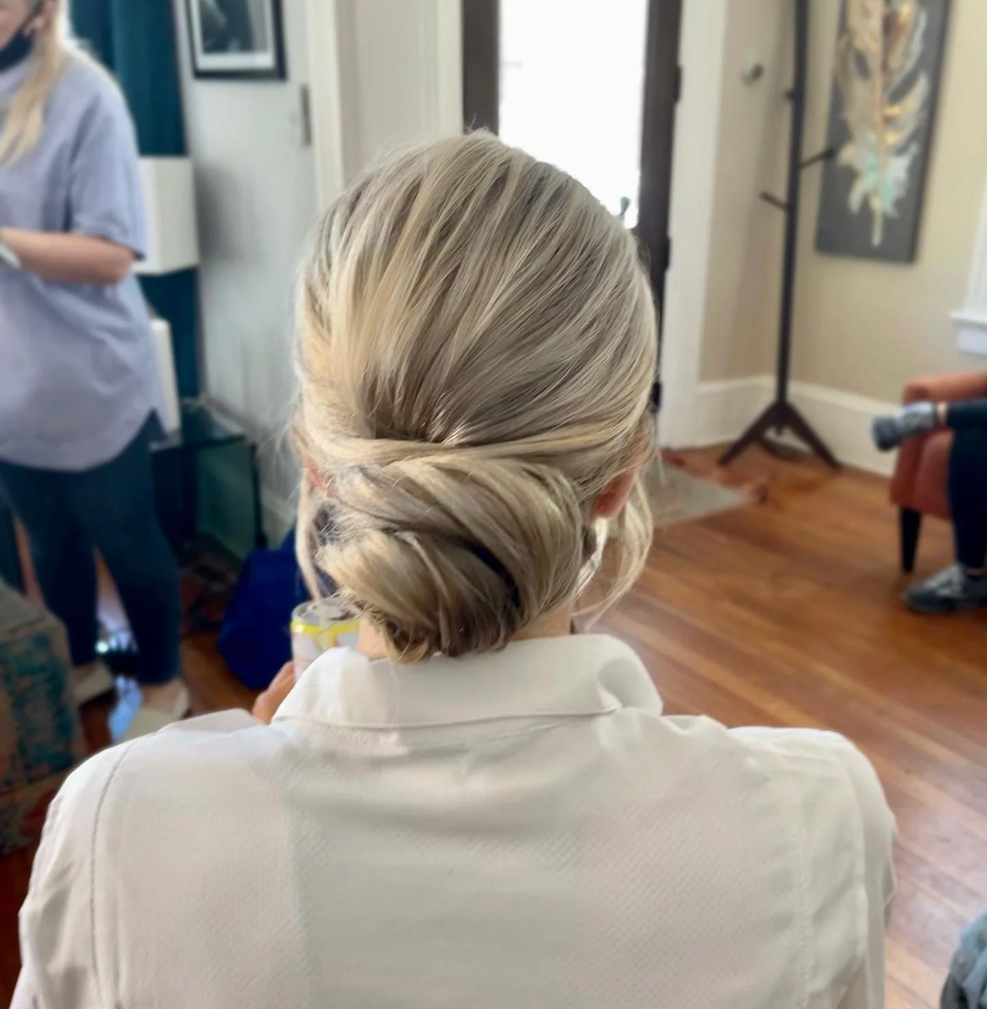 Back view of a woman with blonde hair styled in an elegant updo, wearing a white shirt, in a warmly lit room with wooden floors and a couch.