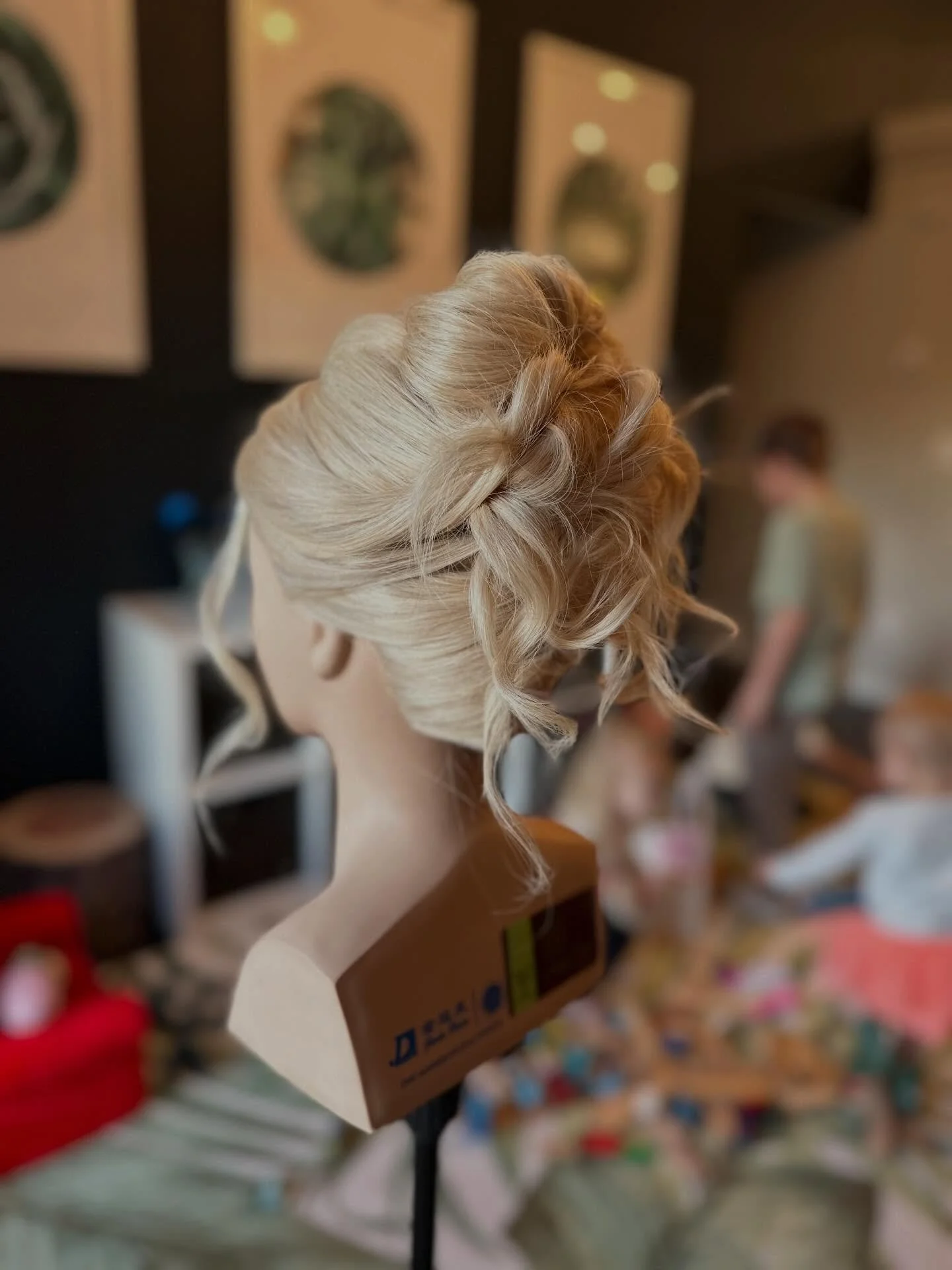 Blonde mannequin head with an elegant updo hairstyle on a salon stand, blurred background with framed pictures and a person in a salon setting.
