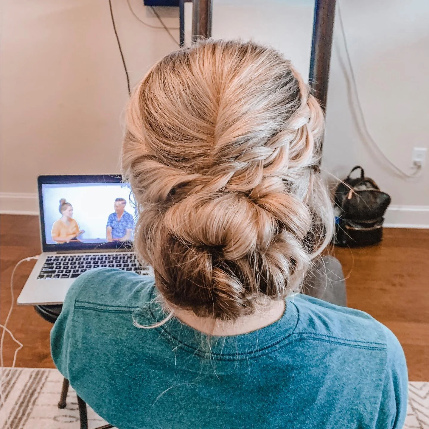 Back view of a woman with blonde hair styled in a loose braided updo, sitting in front of a laptop showing two people on a video call.