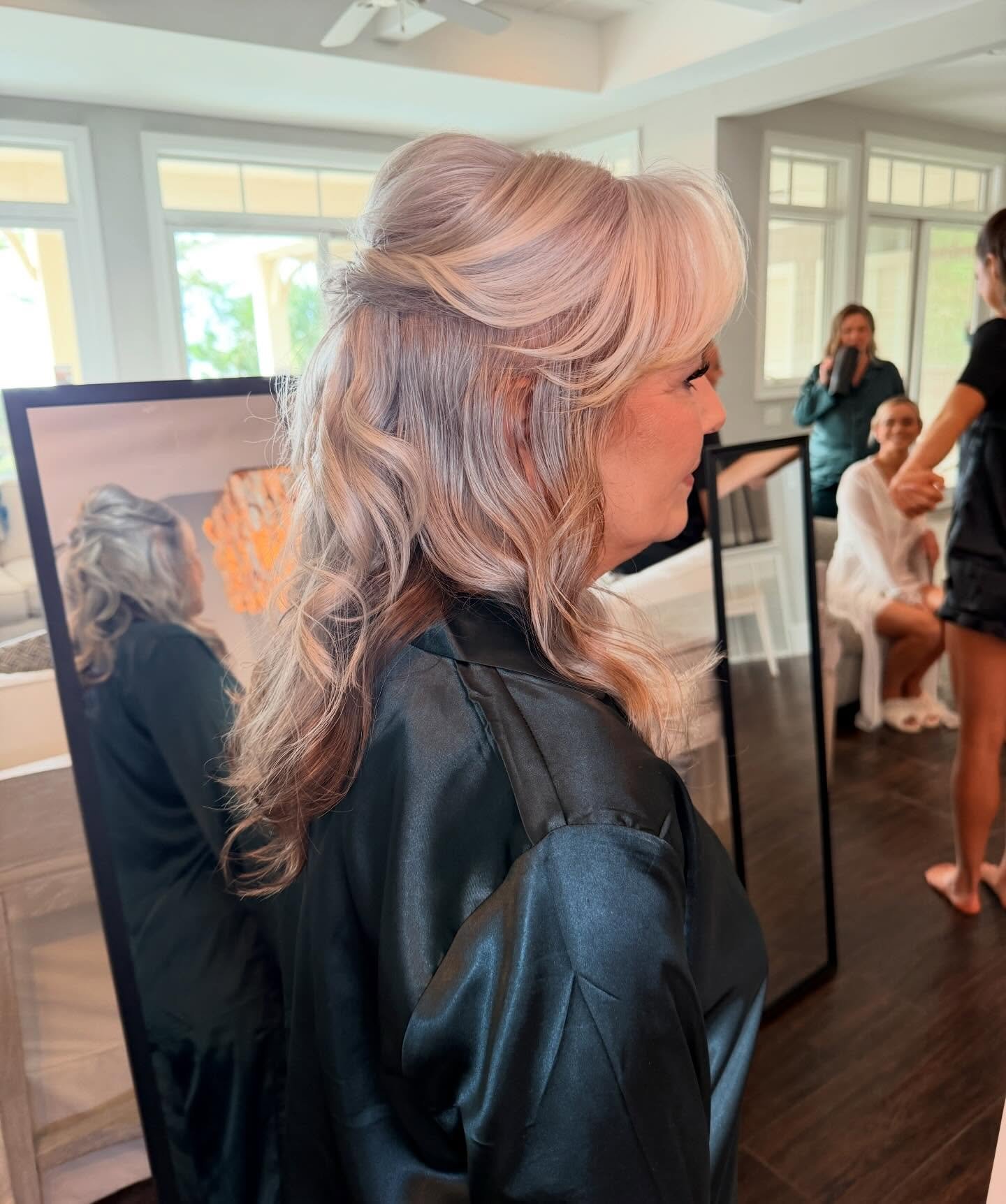 A woman with light blonde, wavy hair wearing a black leather jacket, standing indoors near a mirror, with several people in the background.