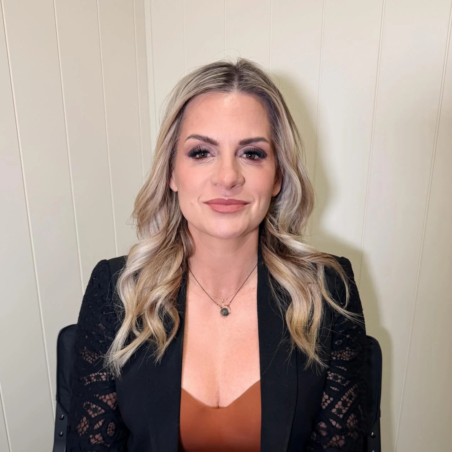 A woman with wavy blonde hair and light skin, wearing makeup, a black jacket with lace sleeves, a brown top, and a black pendant necklace, sitting against a beige wall.