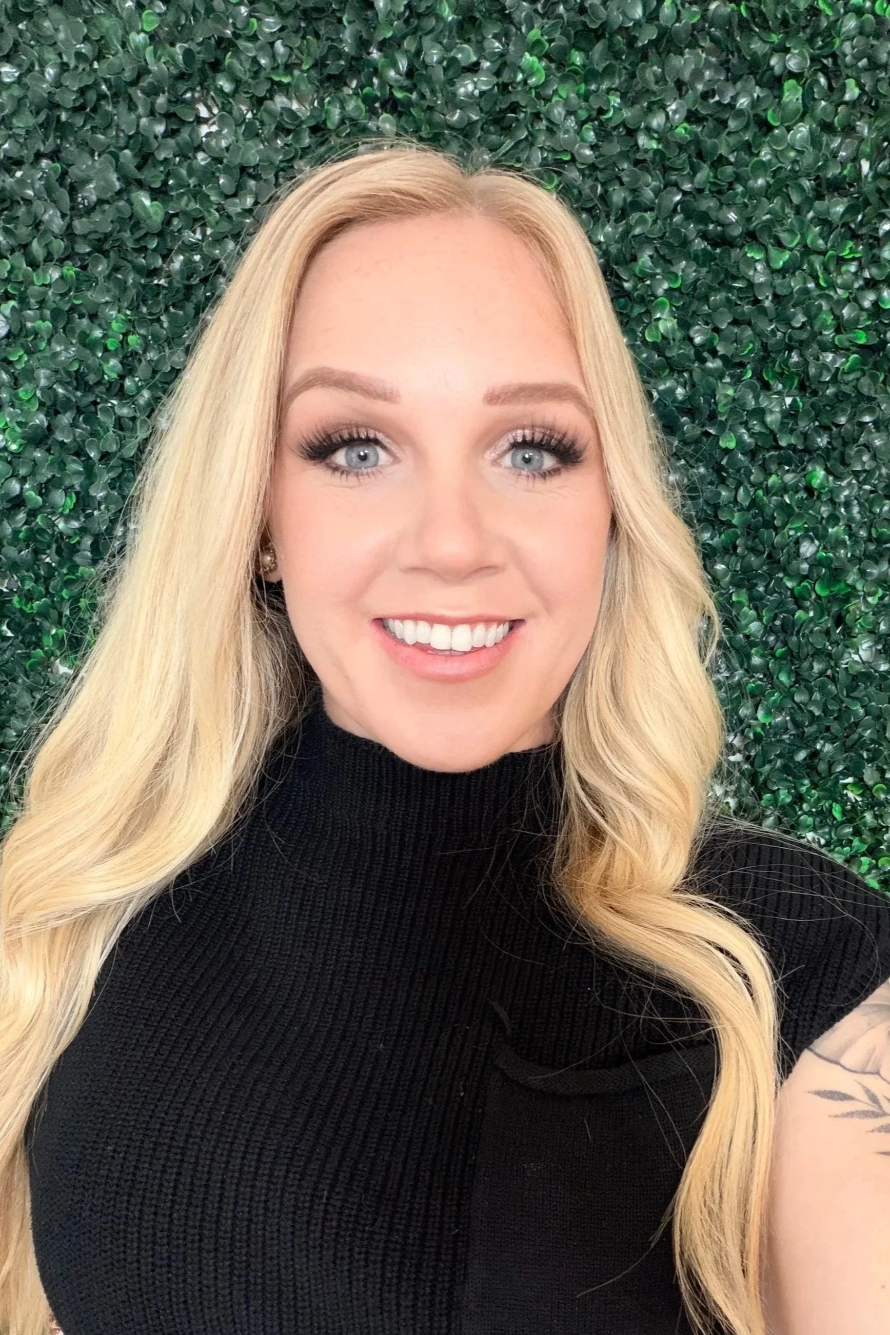 Portrait of a young woman with long blonde hair, wearing a black turtleneck, smiling in front of a green leafy background.