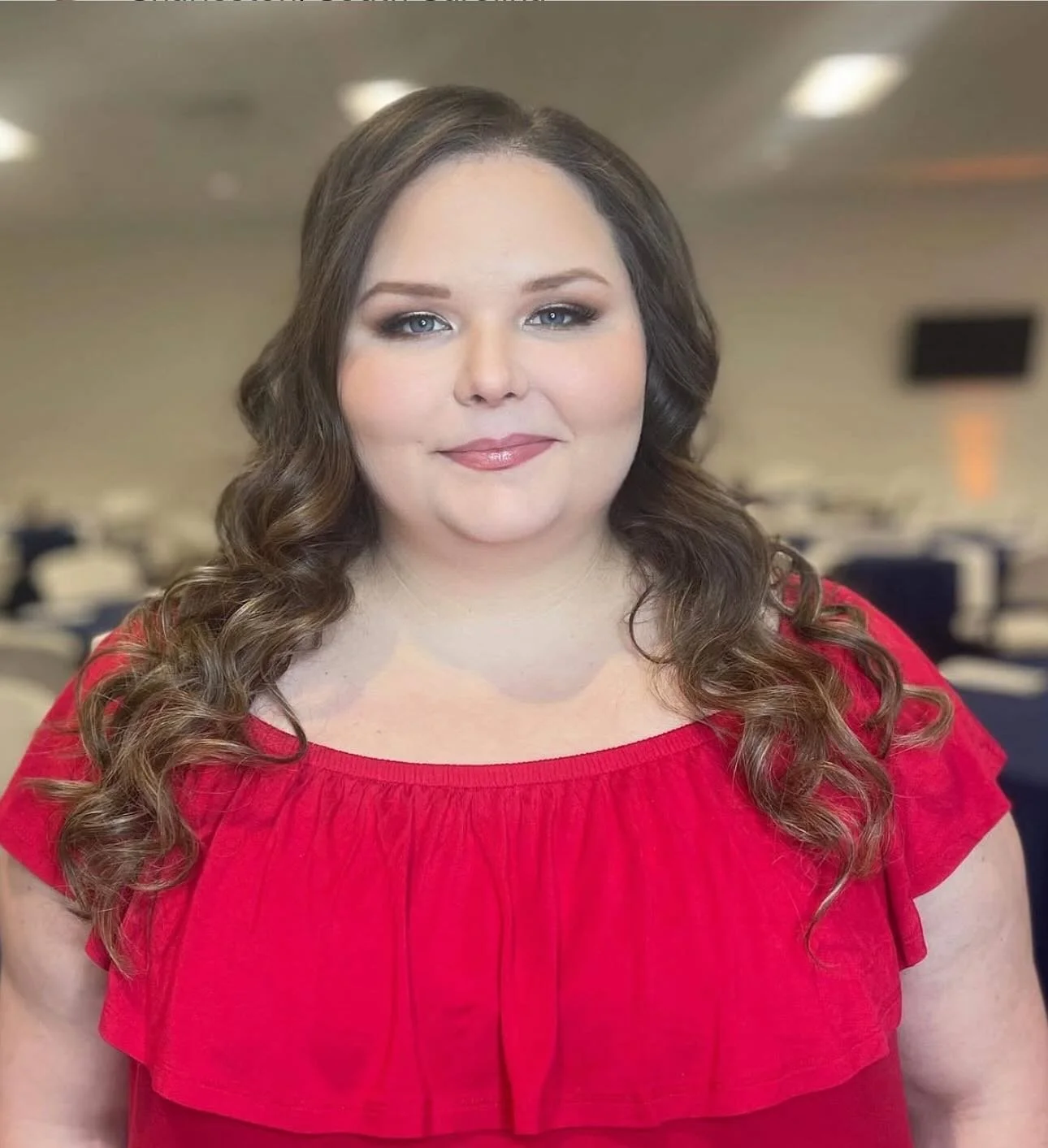 A woman with long, curly brown hair wearing a red top with ruffled neckline, standing in a room with blurred background.