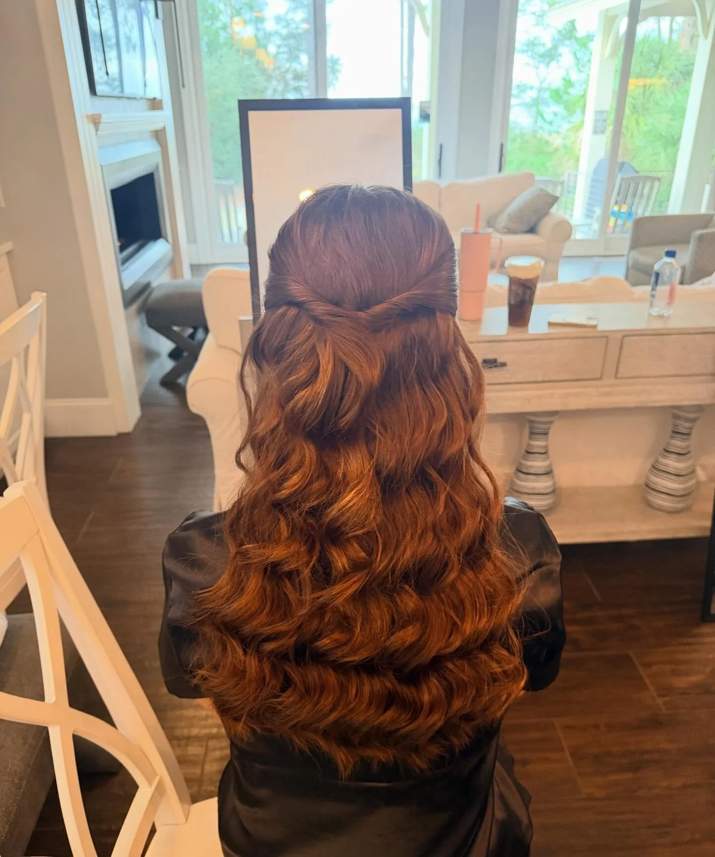 Back of a woman with long, wavy, auburn hair standing in a living room near a window, with drinks and a white sofa in the background.