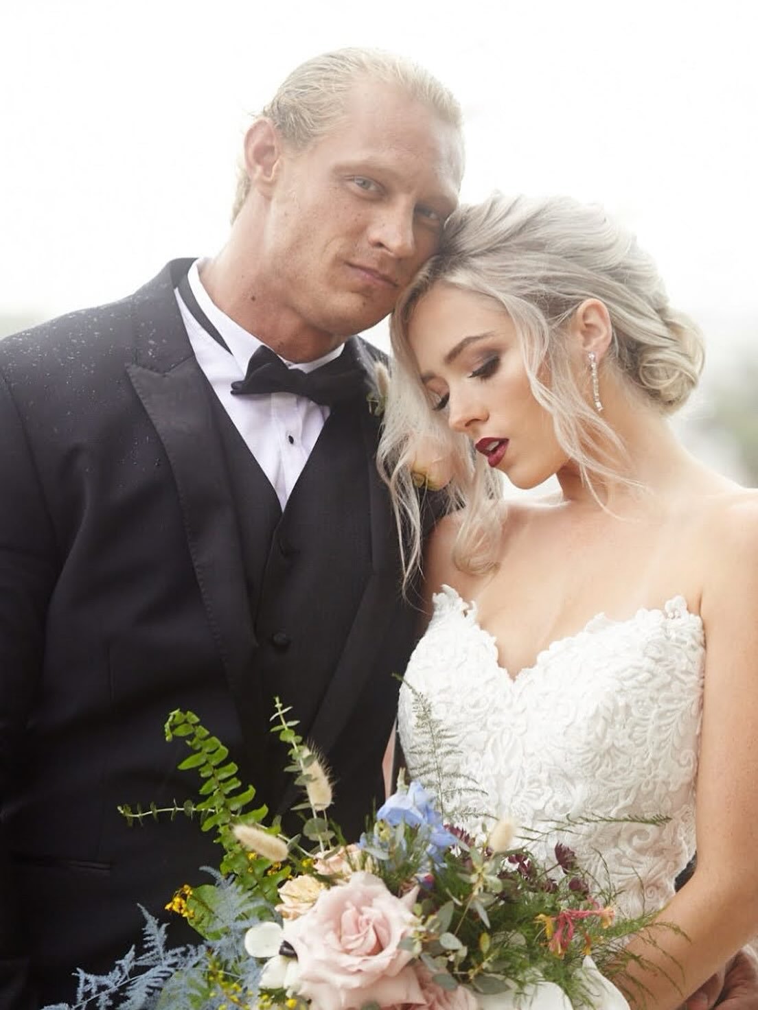 A bride and groom are posing closely together outdoors, with the groom in a black tuxedo and the bride in a white lace wedding dress holding a bouquet of flowers.