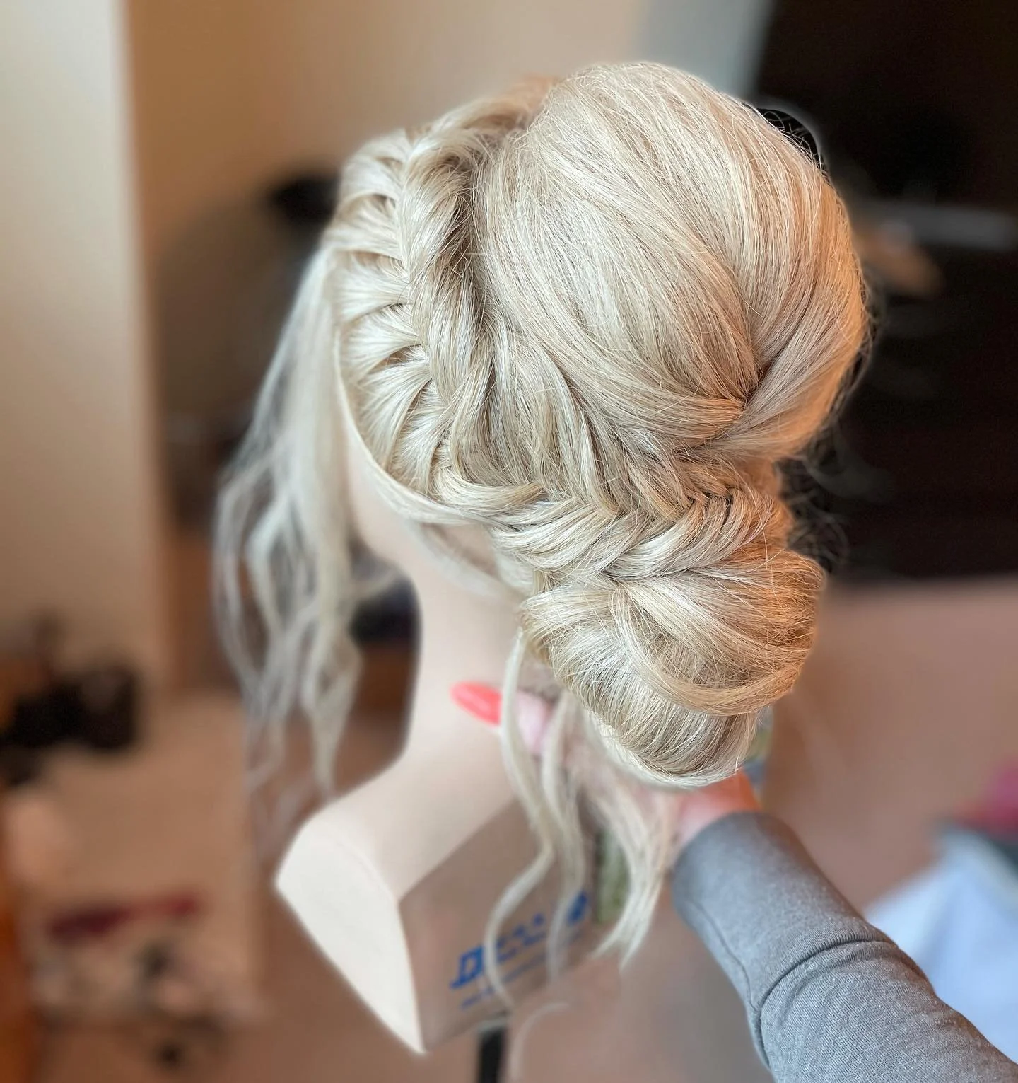 A mannequin head with platinum blonde hair styled in an elegant twisted updo with loose curls, held by a person in a gray long sleeve shirt.