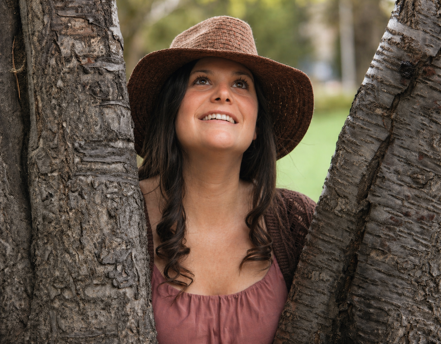 Lauraine Mann, psychosomatic practitioner, smiling up between two trees in a brown hat