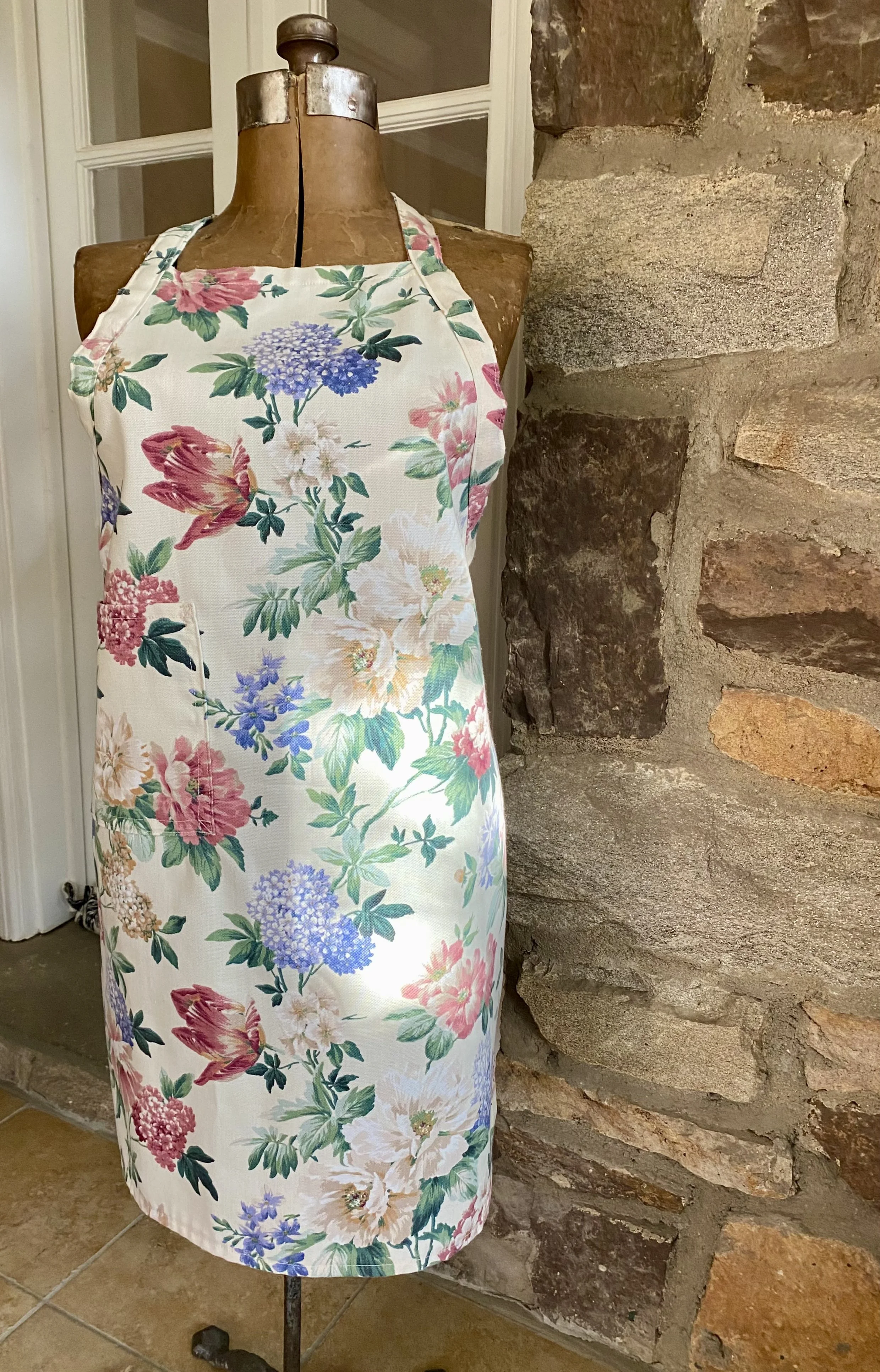 A vintage dress form mannequin wearing a floral apron with pink, purple, white, and green flowers, placed next to a stone wall.