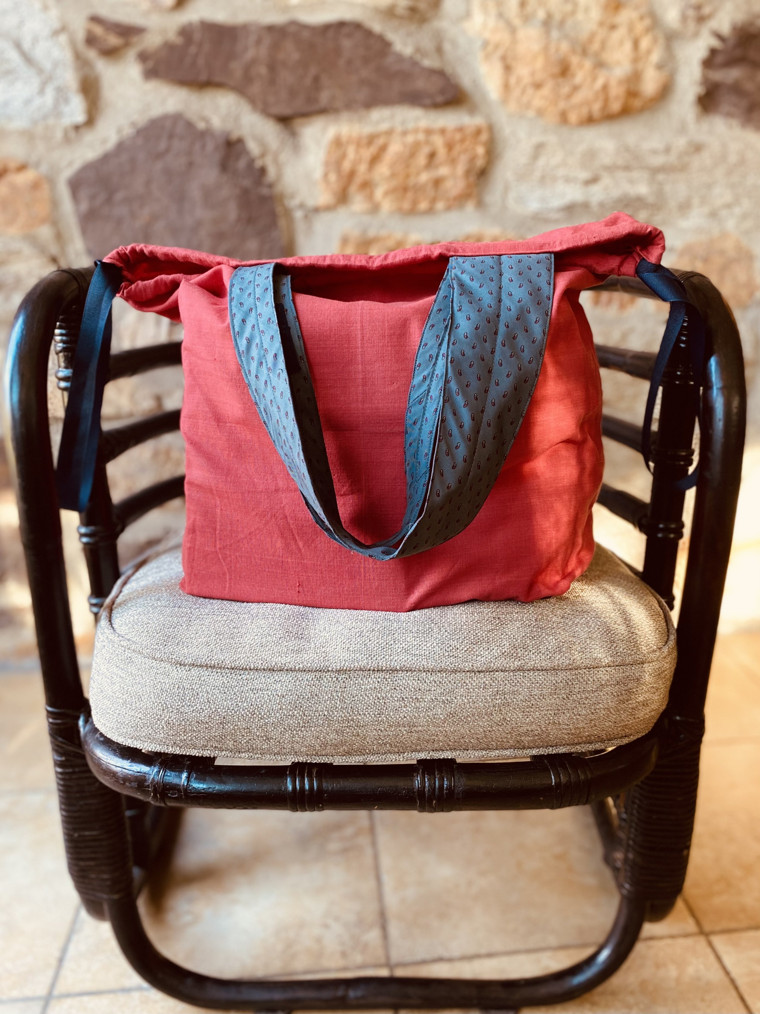 A red tote bag with blue patterned handles resting on a beige cushion on a dark wooden chair against a brick wall background.