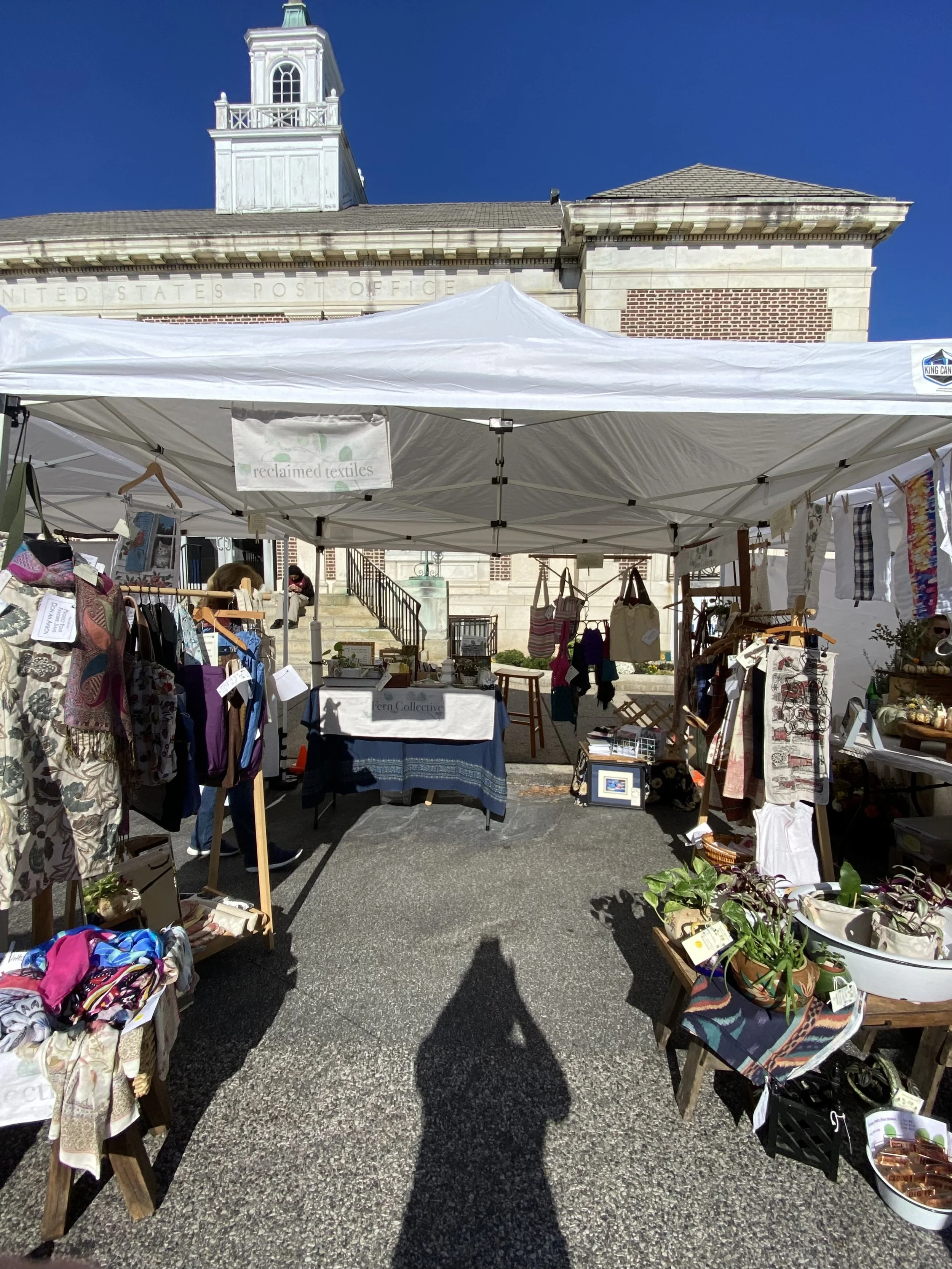 Outdoor market stall with clothing, textiles, and plants in front of a historic building with a steeple, under a clear blue sky.