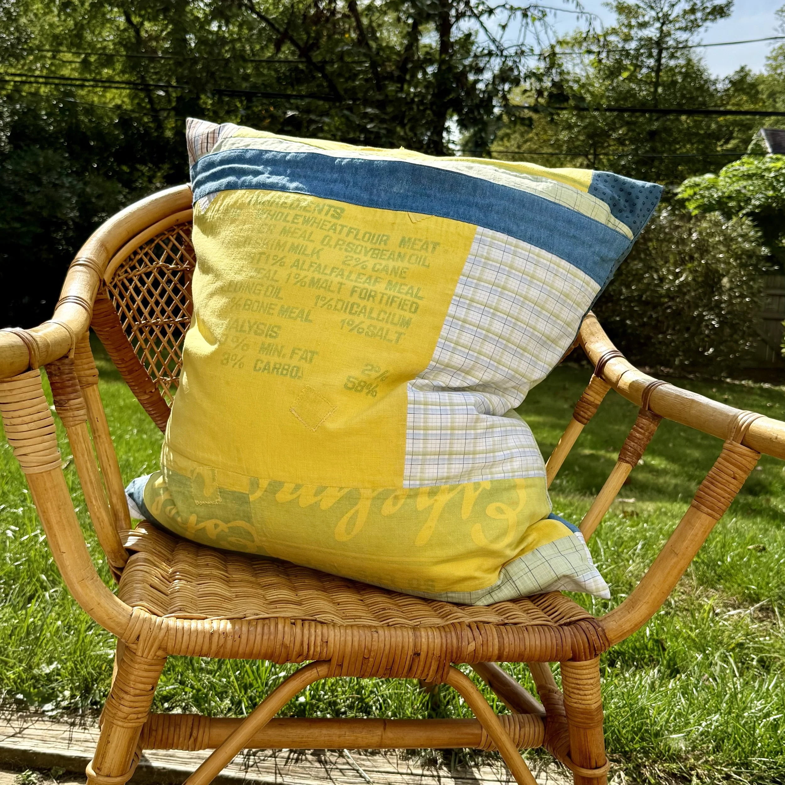 A wicker outdoor chair with a colorful pillow on it, set in a sunny backyard with green grass and trees in the background.