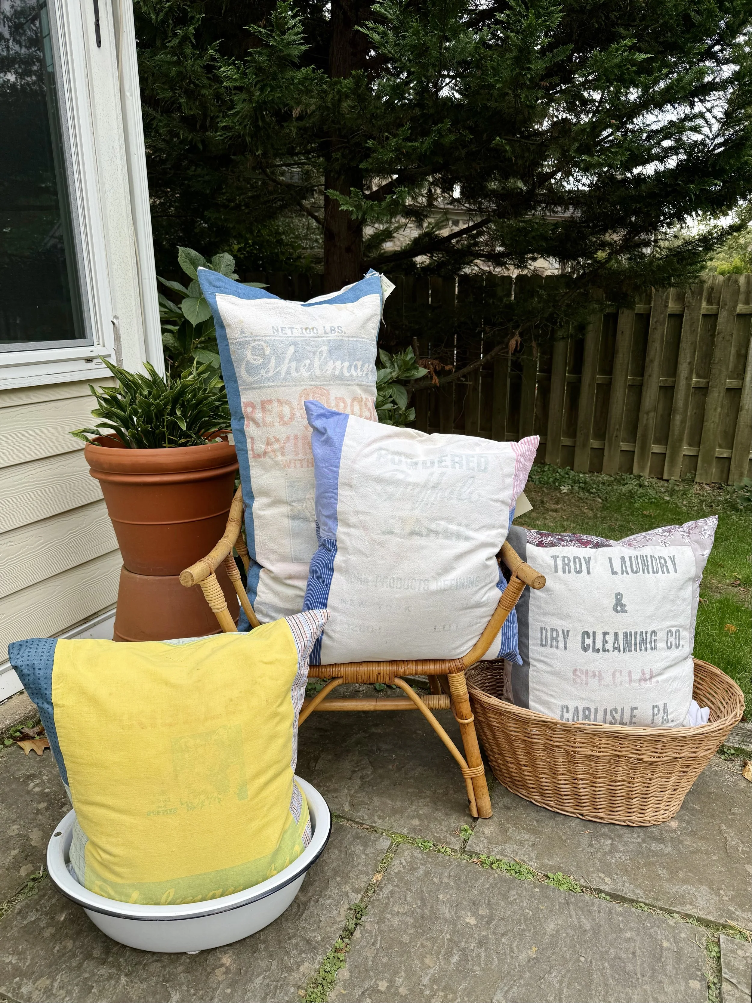Stacked decorative pillows resembling vintage flour and laundry sacks, sitting on a patio with a potted plant, wicker chair, and wooden fence in the background.